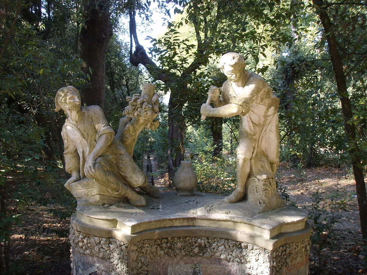 A stone statue depicts two men, one with outstretched arms reclining in a relaxed pose, and the other standing and gesturing, set against a lush forest backdrop in a sun-dappled garden.