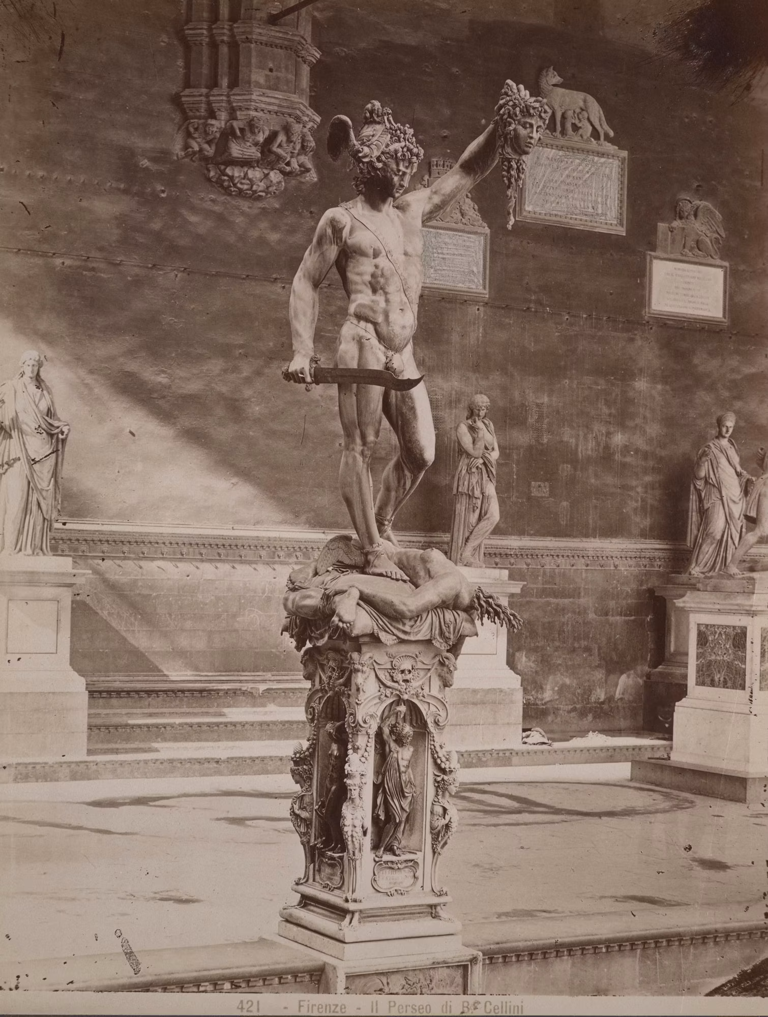 La statua in bronzo raffigura Perseo che solleva la testa di Medusa, situata in un museo con altre statue classiche sullo sfondo.