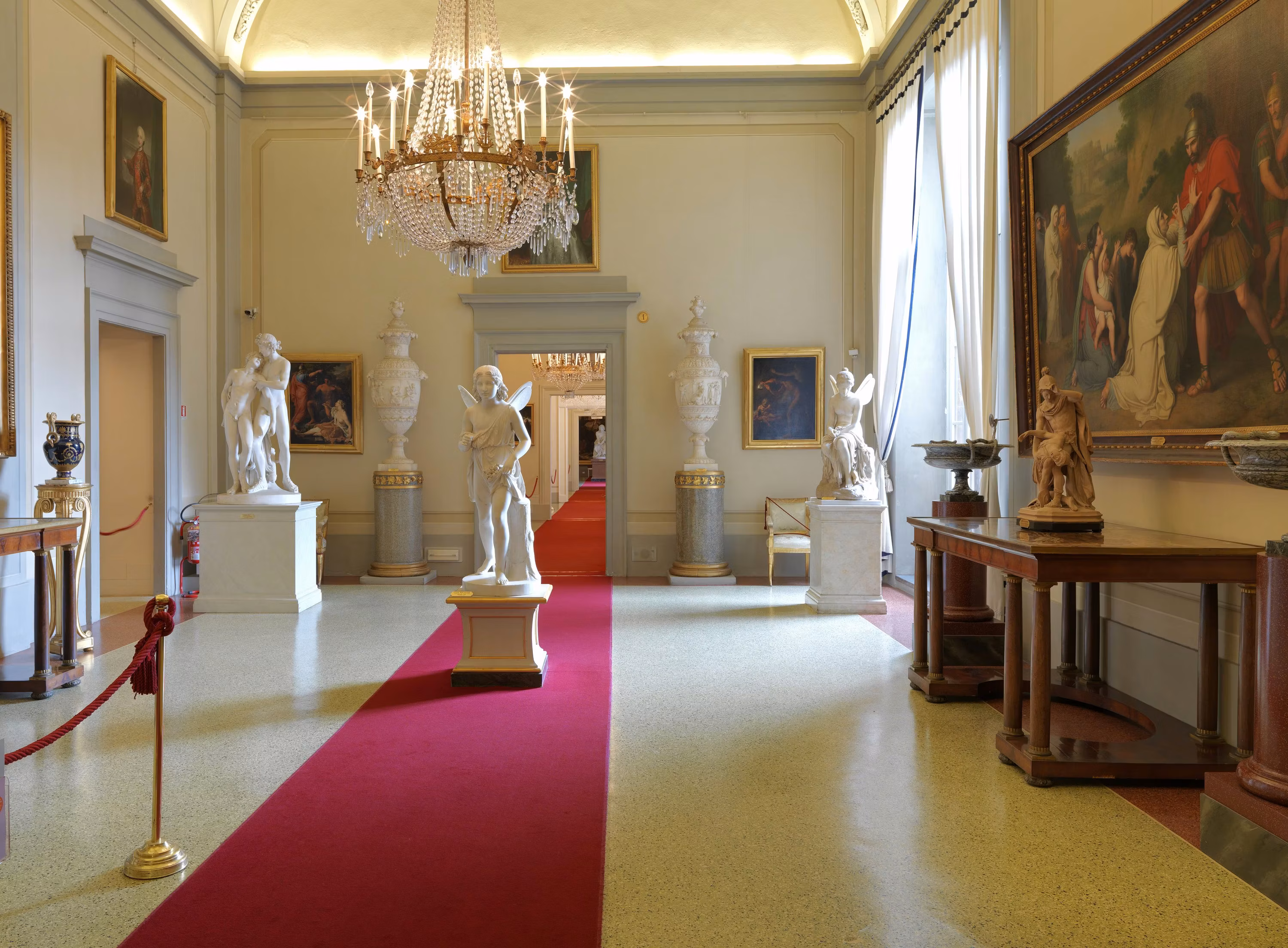 Una lussuosa sala d'arte con un elegante lampadario di cristallo, statue classiche su piedistalli, e un tappeto rosso che percorre il centro dello spazio, con grandi quadri che adornano le pareti.