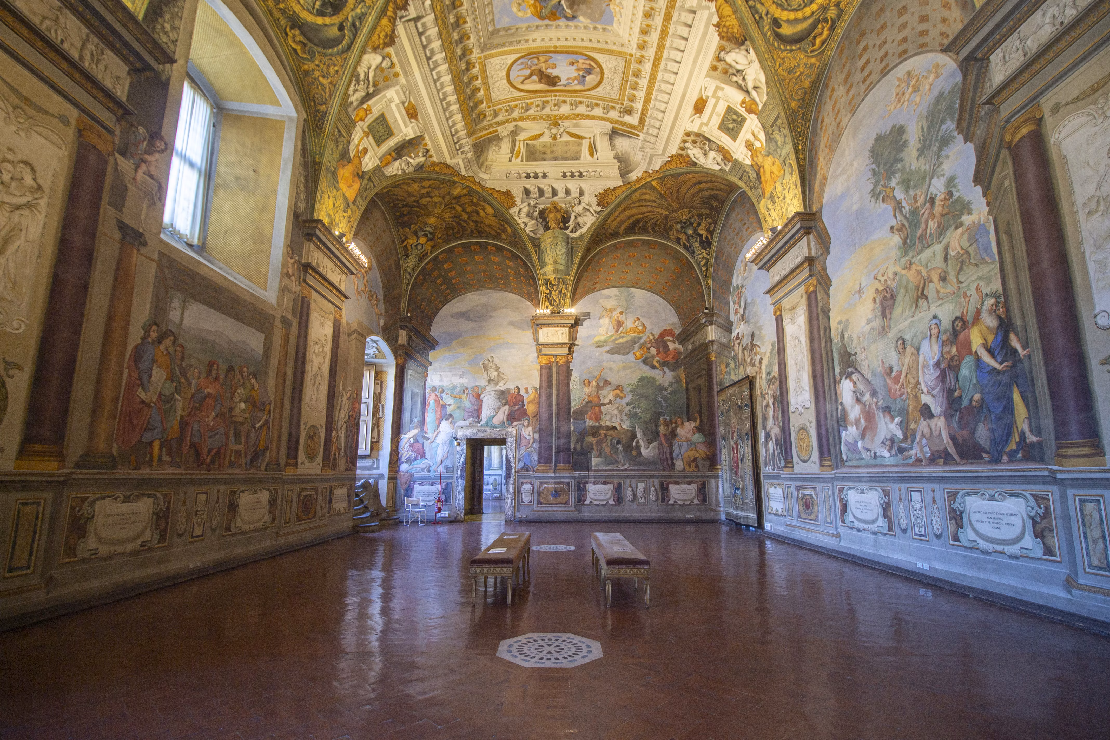A grand hall with intricately painted frescoes covering the tall walls and vaulted ceiling, showcasing vibrant historical and religious scenes, with polished wooden benches on a terracotta-tiled floor.