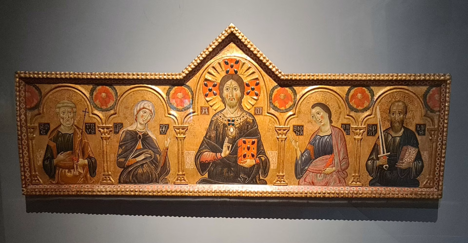 This image features a medieval religious altarpiece with five ornately painted figures, including a central figure with a halo, surrounded by decorative arches and gold accents, displayed in a museum setting.
