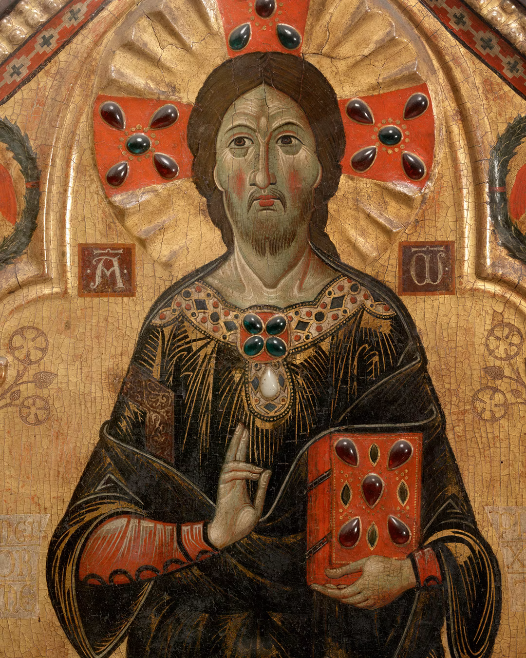 An ornate religious painting depicts a solemn figure with a halo, dressed in richly detailed robes, holding a book adorned with red gemstones, set against a backdrop with intricate red and gold designs.