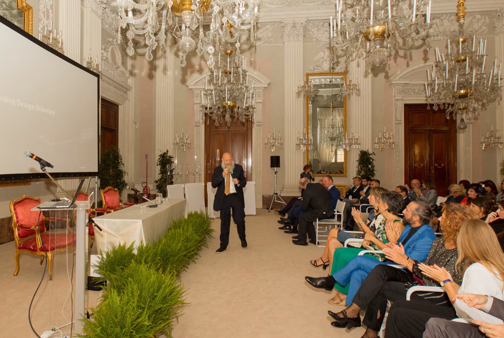 In un'elegante sala ornata da lampadari di cristallo e decorazioni elaborate, un conferenziere parla a un pubblico seduto su sedie bianche, attento alla presentazione che si svolge su uno schermo a sinistra.