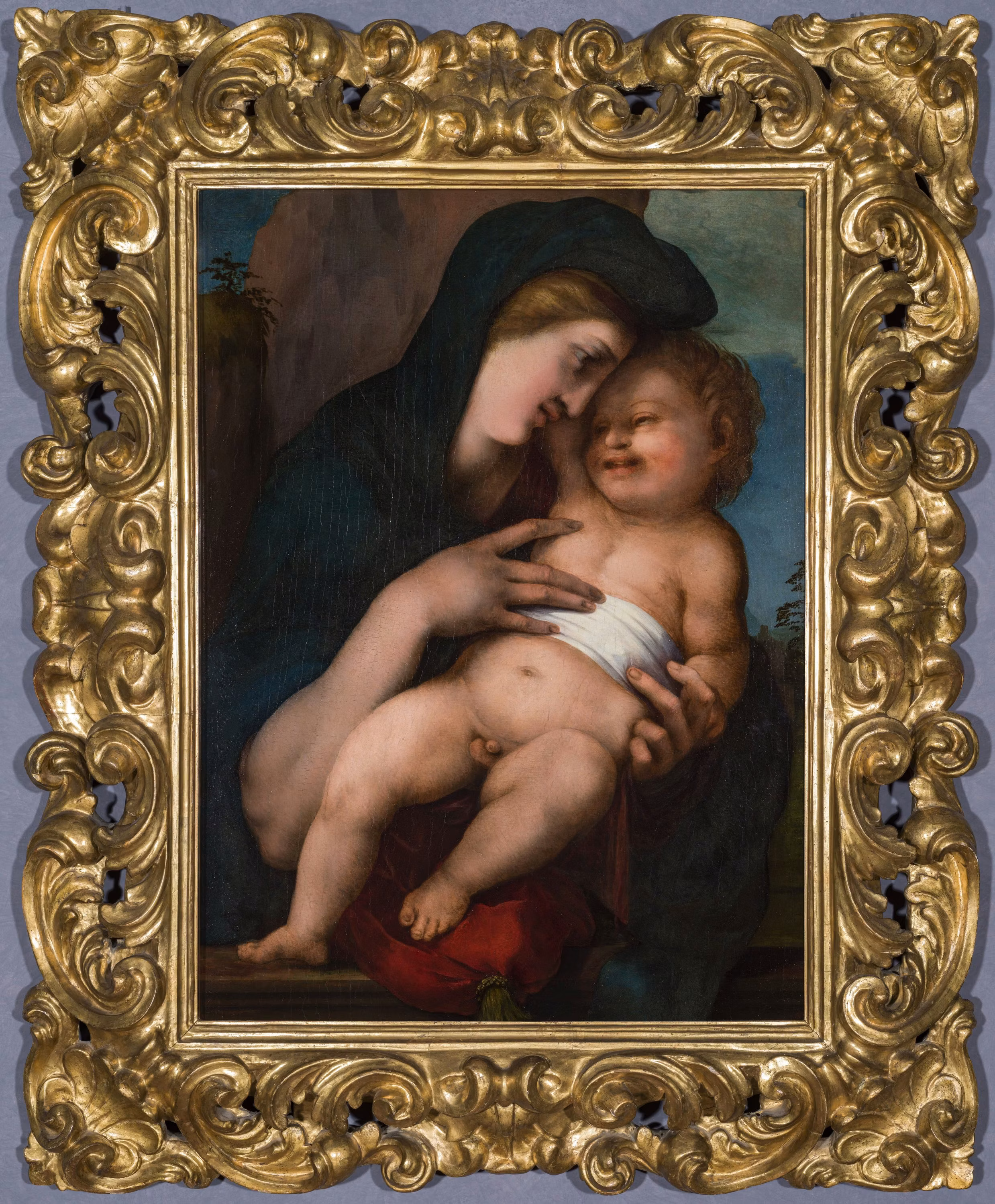 A painting of a mother gently holding her baby, both wearing draped clothing, set within an ornate golden frame.