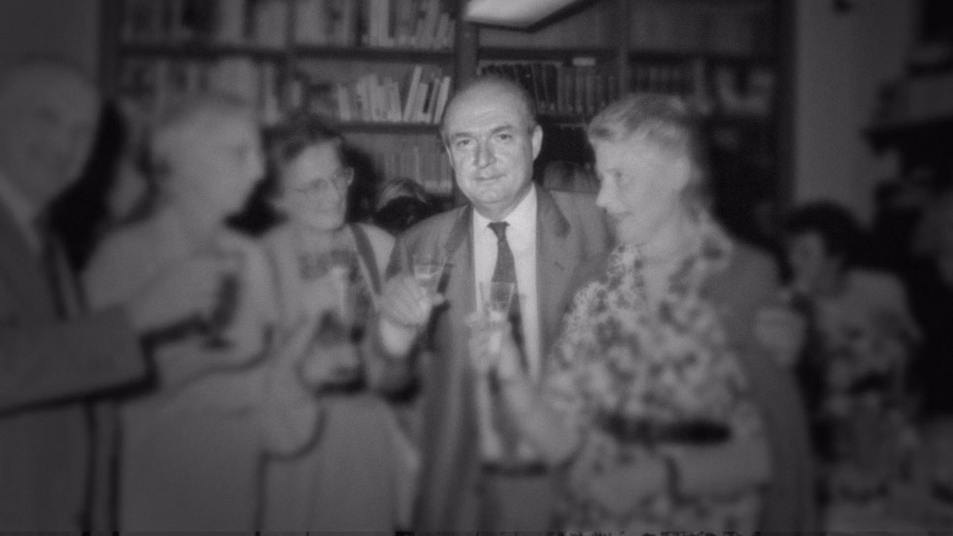 In una sala affollata, un gruppo di persone eleganti brinda con bicchieri di champagne davanti a uno sfondo di scaffali pieni di libri.