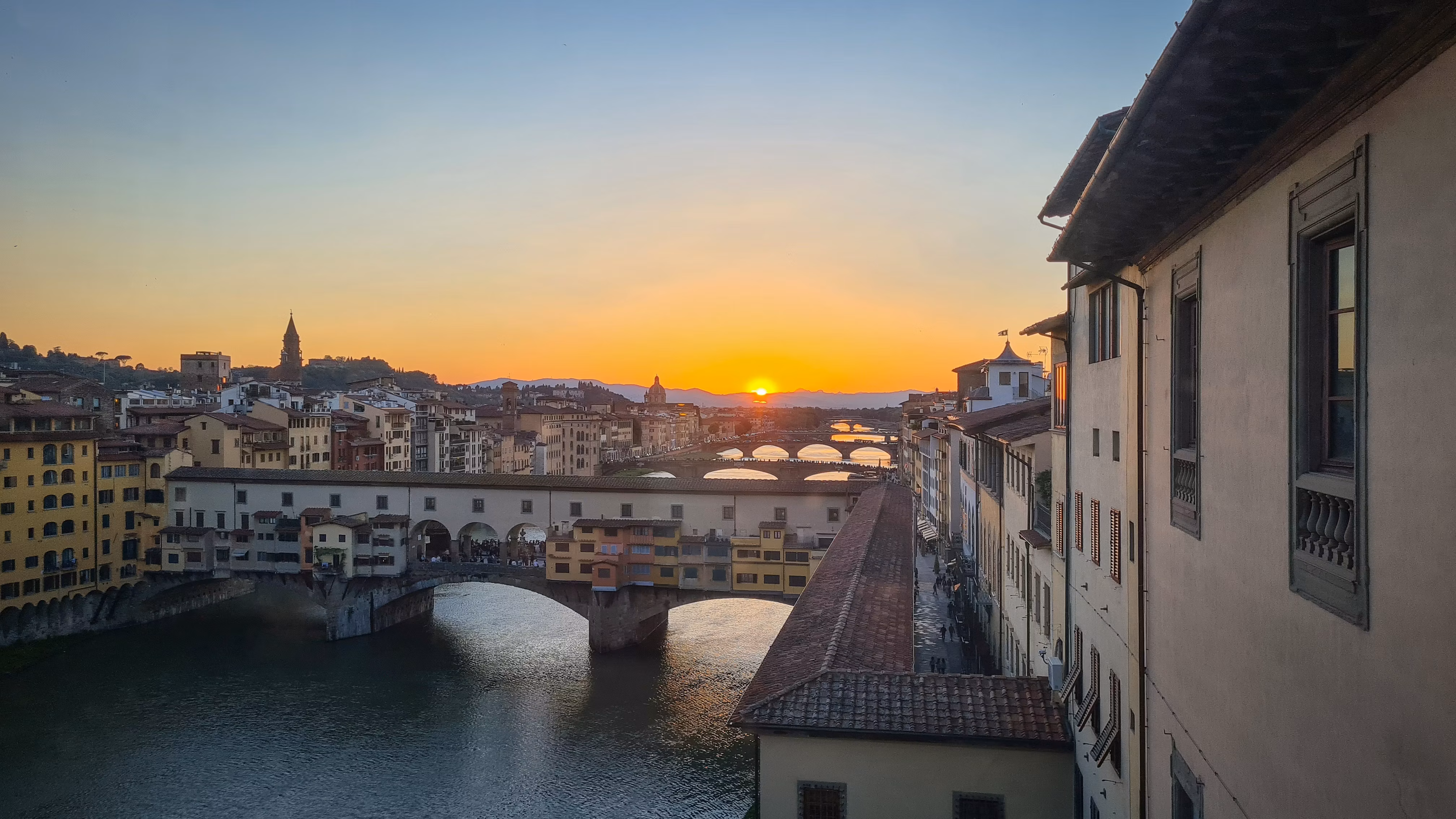 Veduta panoramica del tramonto a Firenze, con il fiume Arno che attraversa la città e il famoso Ponte Vecchio in primo piano, circondato da edifici storici e un cielo sereno e colorato.