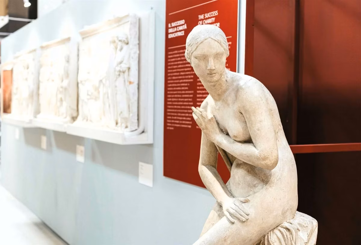 Una statua marmorea di una figura femminile seduta è posizionata in primo piano, con rilievi dettagliati e pannelli informativi esposti su uno sfondo dai toni chiari e rossi in un ambiente museale.