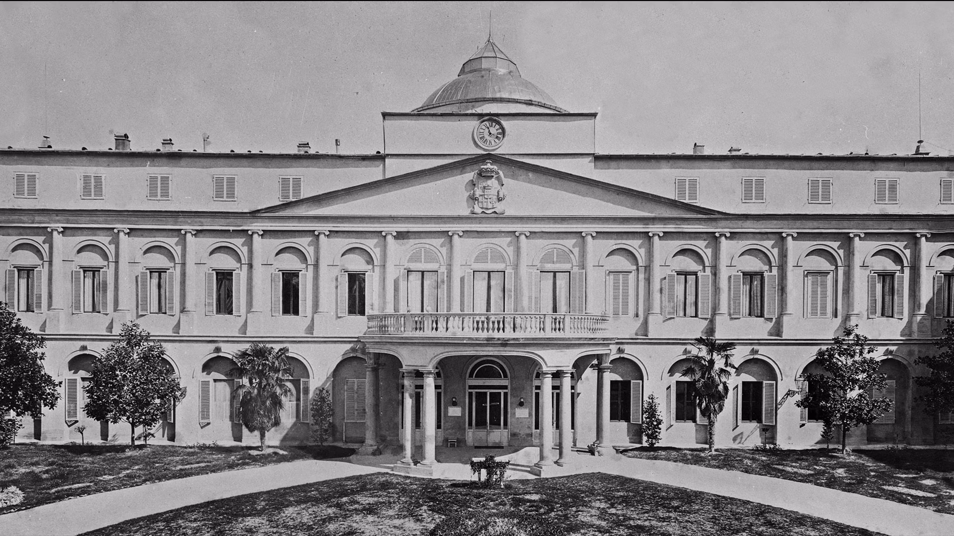 A historic building with a symmetrical facade features arched windows and a prominent central clock above its entrance, surrounded by manicured gardens and a path leading to the entrance.
