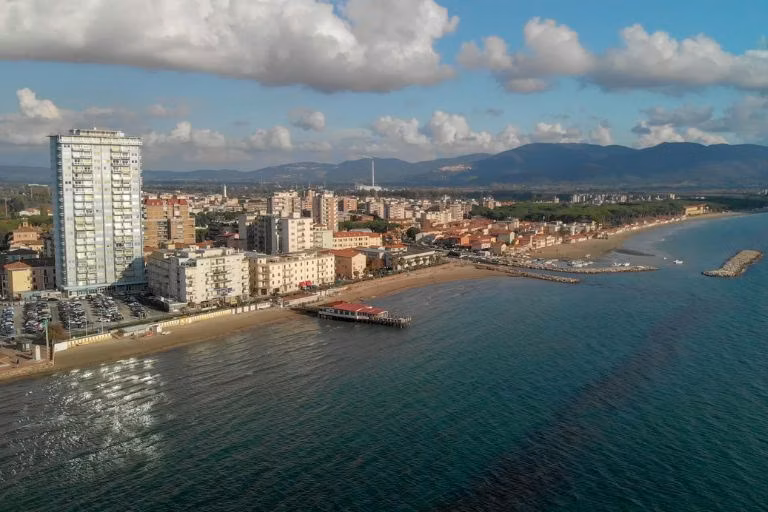Aerial view of a coastal cityscape with a prominent high-rise building near the shoreline, sandy beaches, and a scenic backdrop of mountains under a partly cloudy sky.
