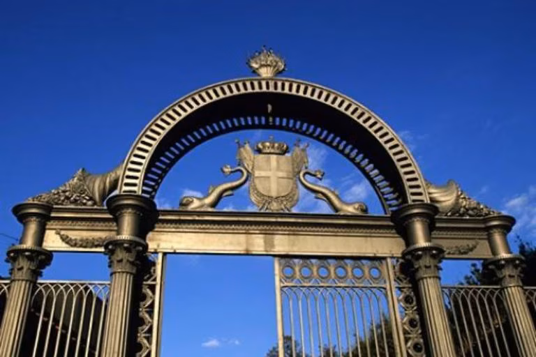 A majestic wrought iron gate stands against a clear blue sky, featuring intricate designs, a decorative arch, and a central coat of arms with heraldic symbols, exemplifying classic architectural elegance.