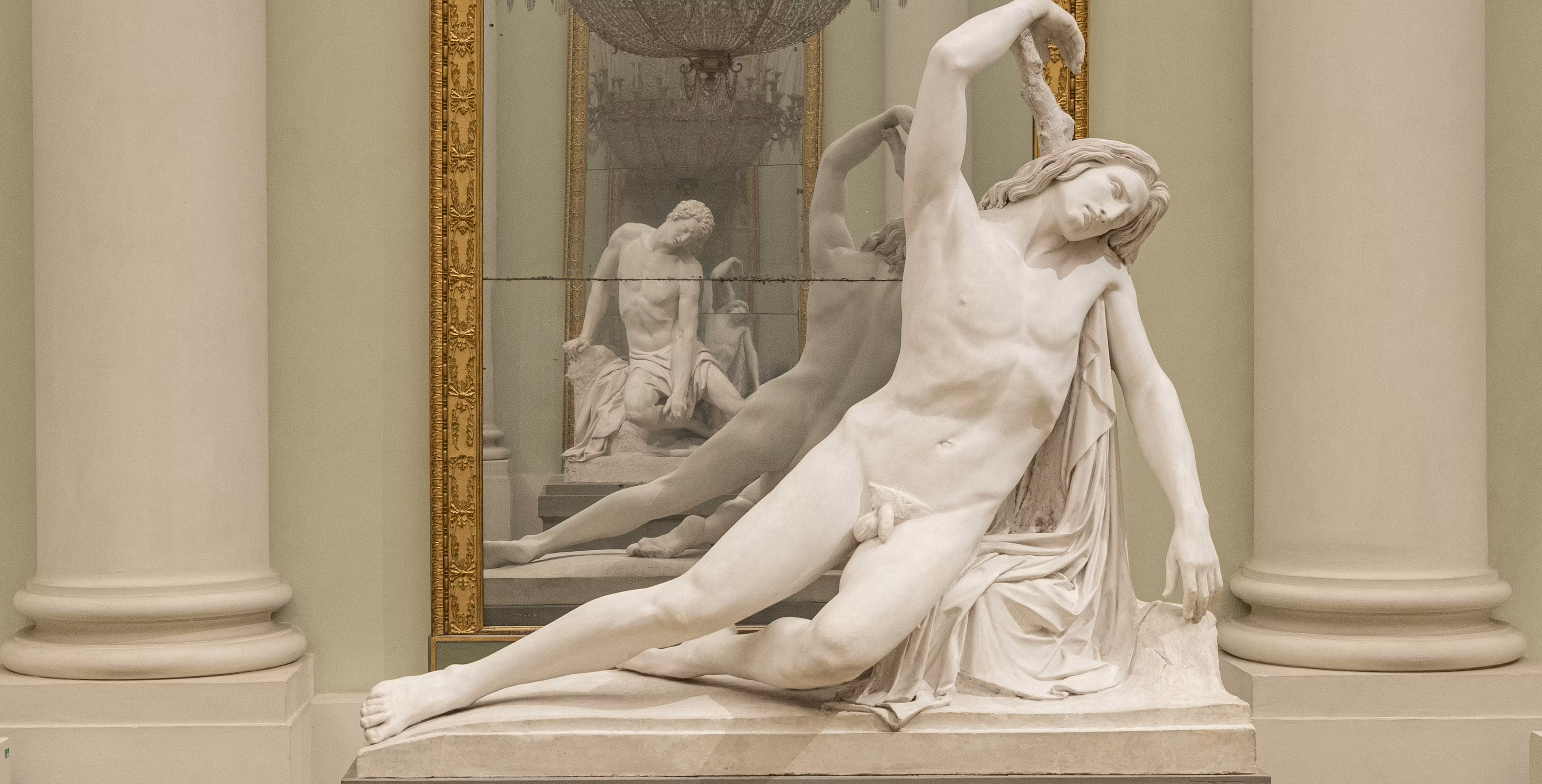 Una statua classica di un corpo umano disteso si riflette in uno specchio dorato tra colonne ornate, con dettagli accurati che mettono in risalto l'anatomia e l'espressione artistica.