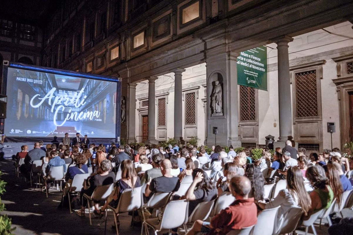 Persone sedute in un cortile notturno, davanti a uno schermo gigante che proietta un film sotto un cielo sereno durante un evento di cinema all'aperto.