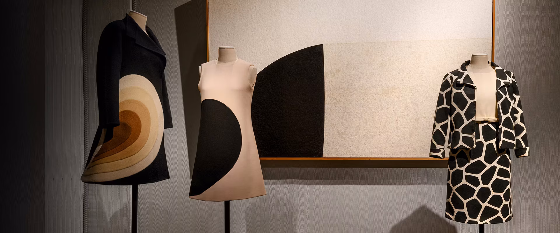 Tre manichini esibiscono abiti vintage con design geometrici audaci di diverse tonalità di nero, bianco e marrone, posizionati davanti a un grande quadro astratto che riflette motivi simili.