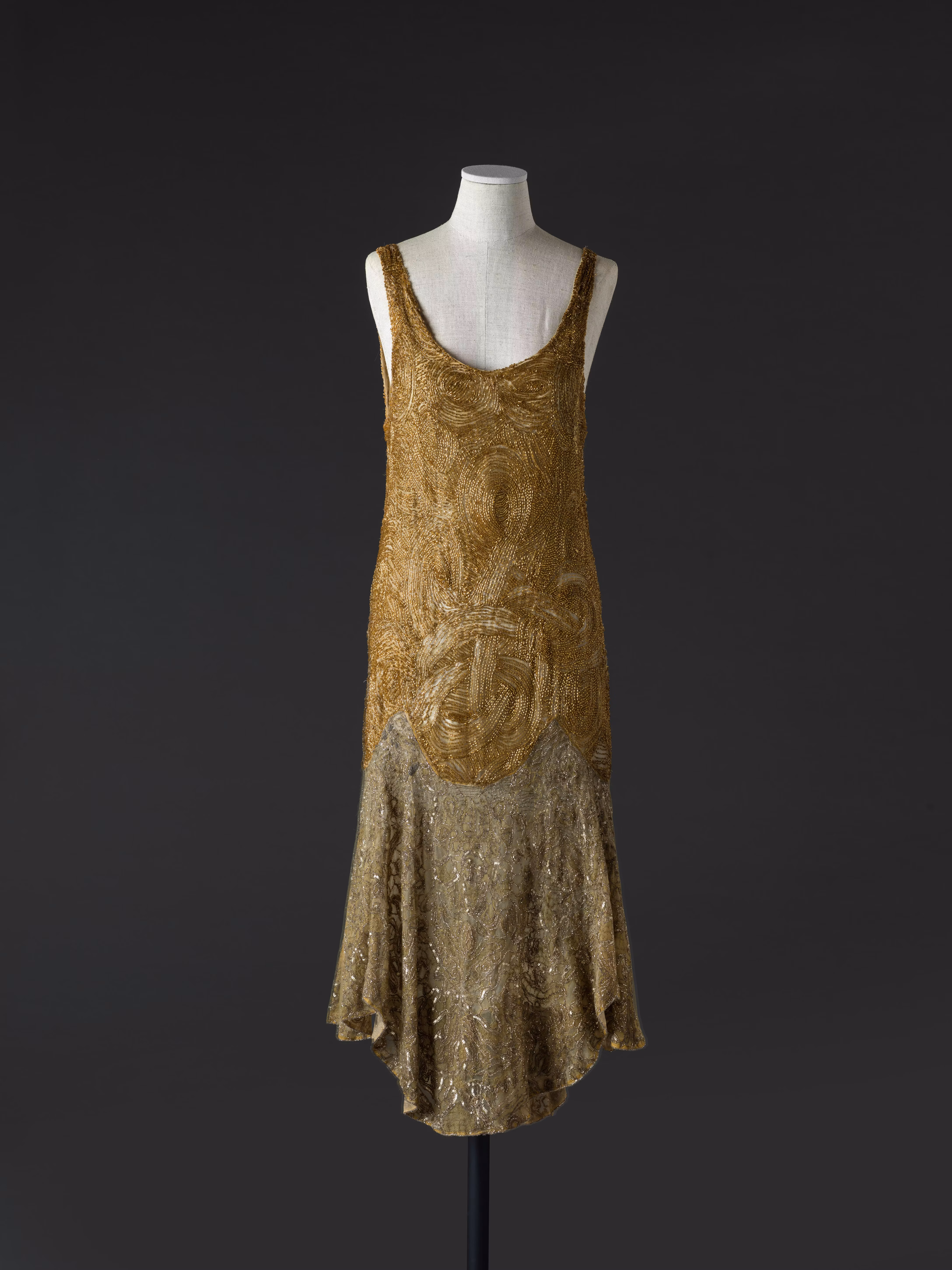 An elegant, sleeveless flapper dress from the 1920s, featuring luxurious gold and silver beading in intricate patterns, displayed on a mannequin against a dark background.