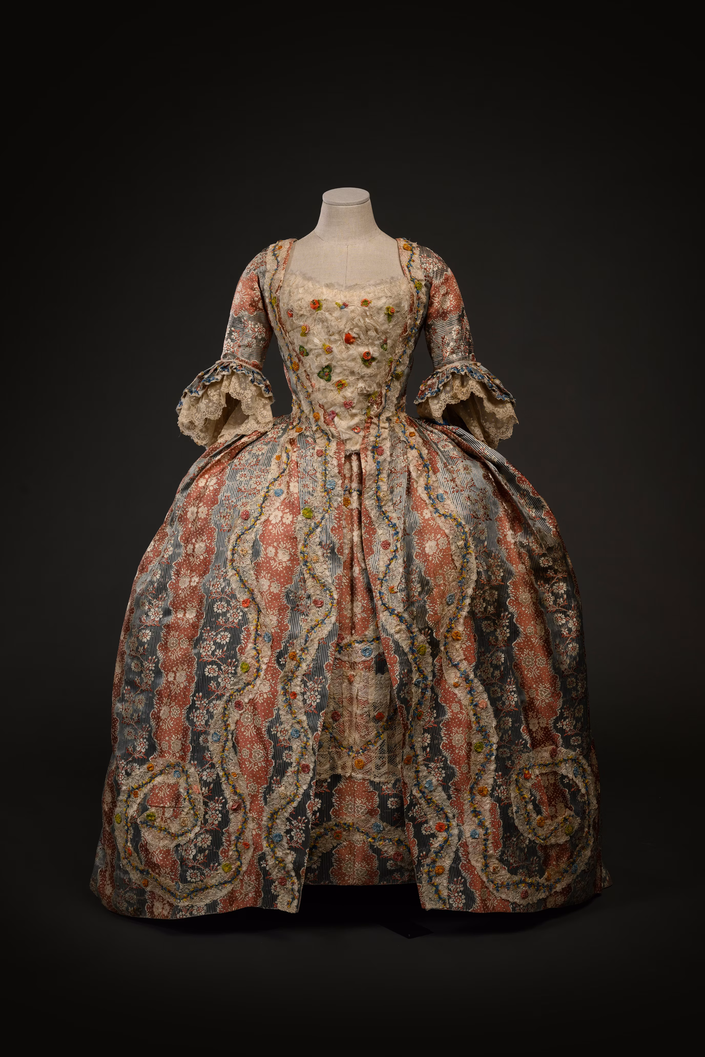An elegant 18th-century gown displayed on a mannequin, featuring intricate floral patterns in muted tones and a voluminous skirt, exemplifying historical fashion design.
