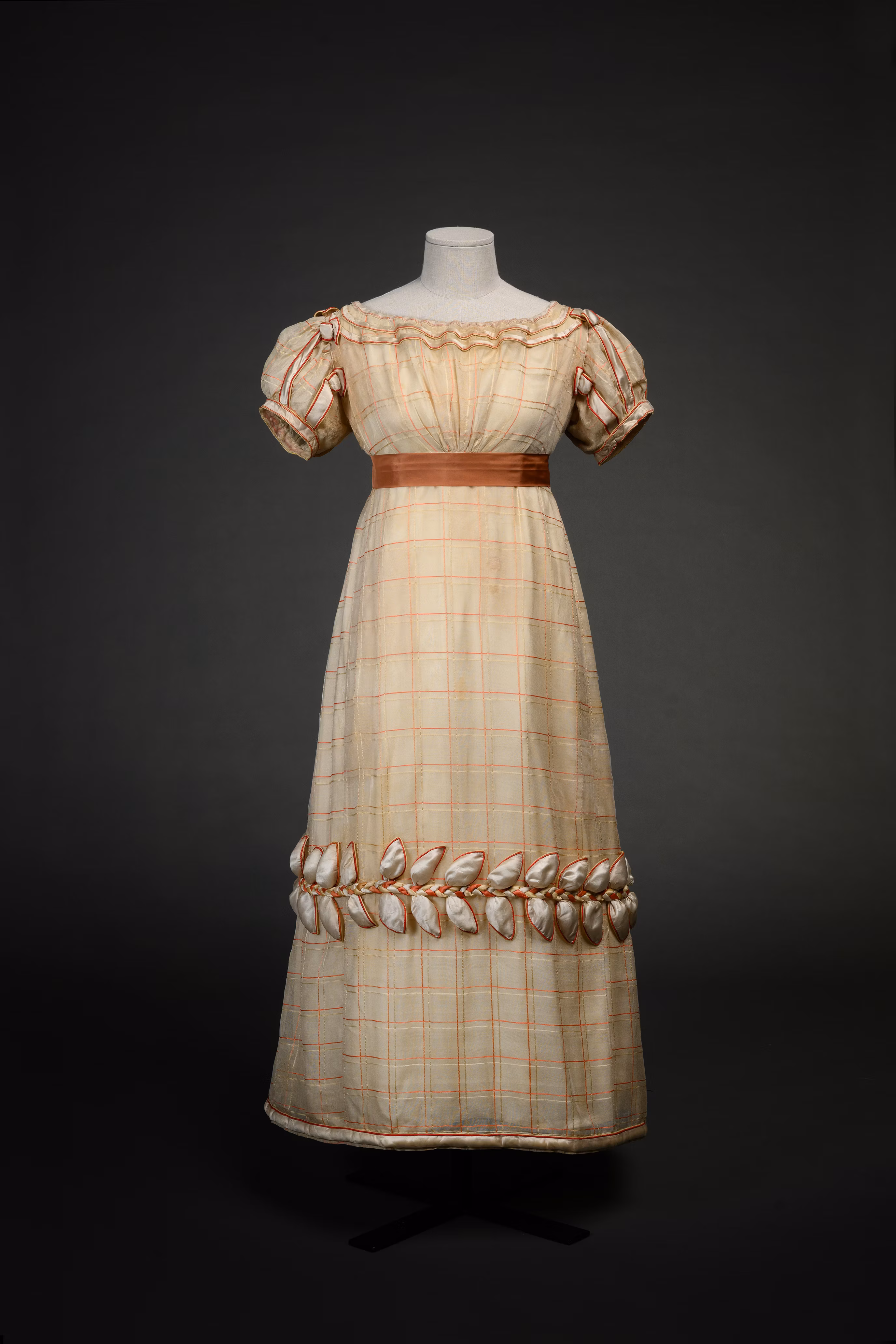 A vintage, Regency-style dress is displayed on a mannequin, featuring short puff sleeves, a high waist with a ribbon belt, and a checkered pattern accented with ribbon detailing along the hem.