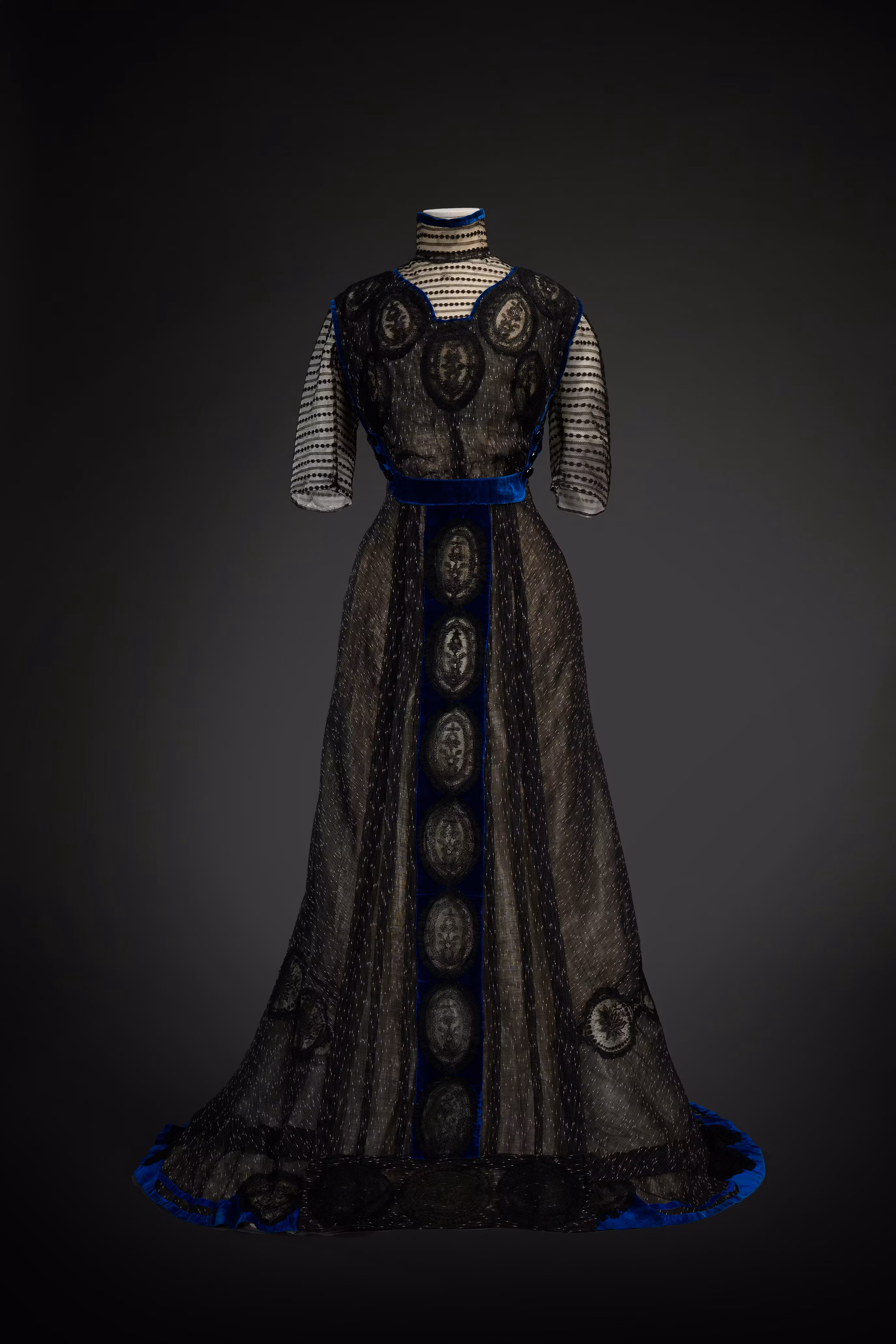 A vintage black evening gown with intricate lace detailing and striking blue velvet accents is displayed elegantly against a dark background, showcasing its full-length silhouette and timeless design.