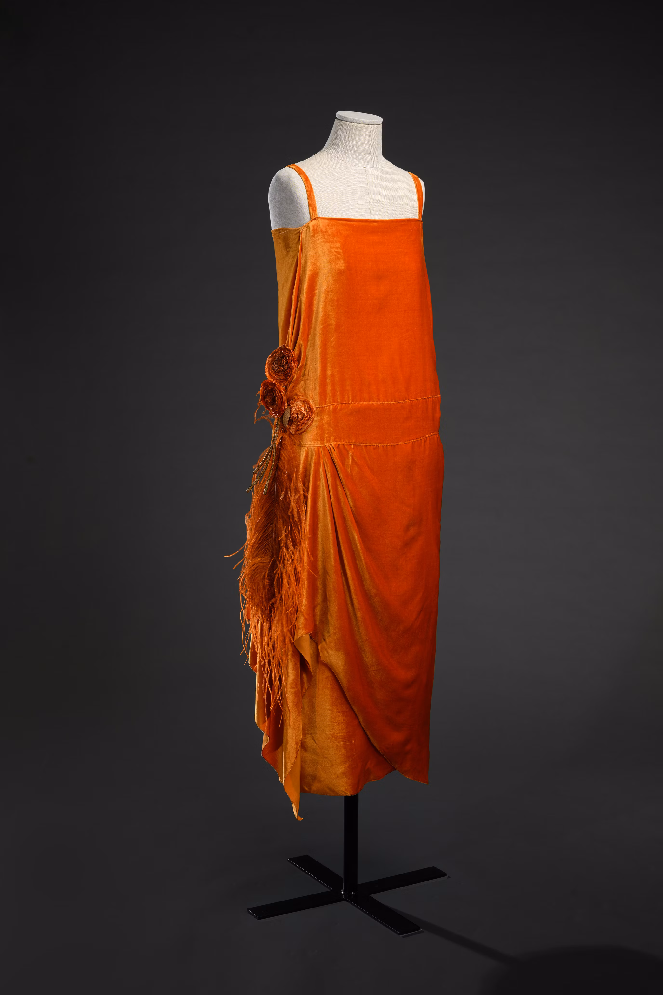 Vestito arancione senza maniche su manichino, con dettagli floreali e frange lungo un lato, visualizzato su sfondo scuro.