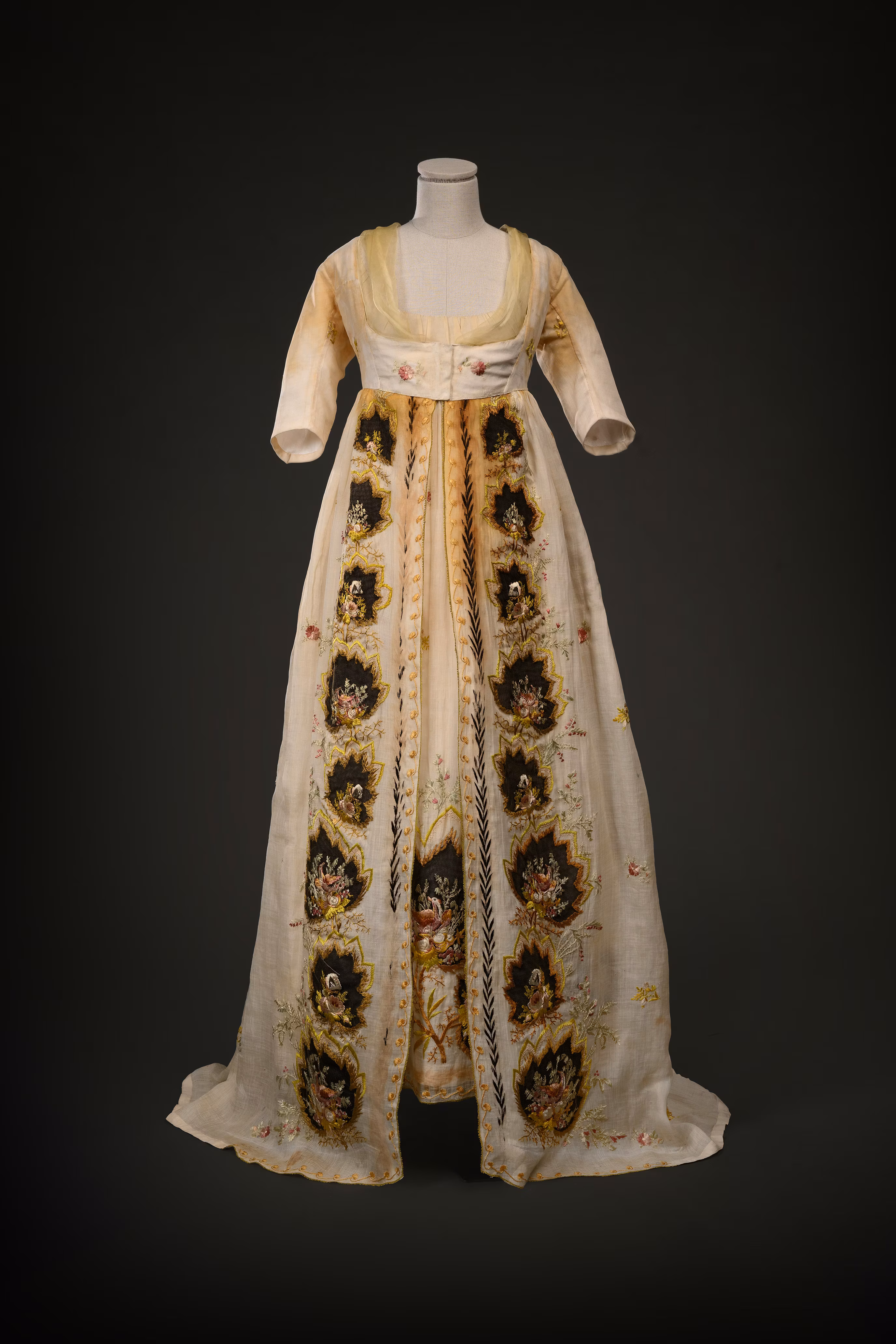 A vintage Regency-era high-waisted gown displayed on a mannequin, featuring intricate floral embroidery on a cream-colored fabric with a sheer overlay, highlighted against a dark background.