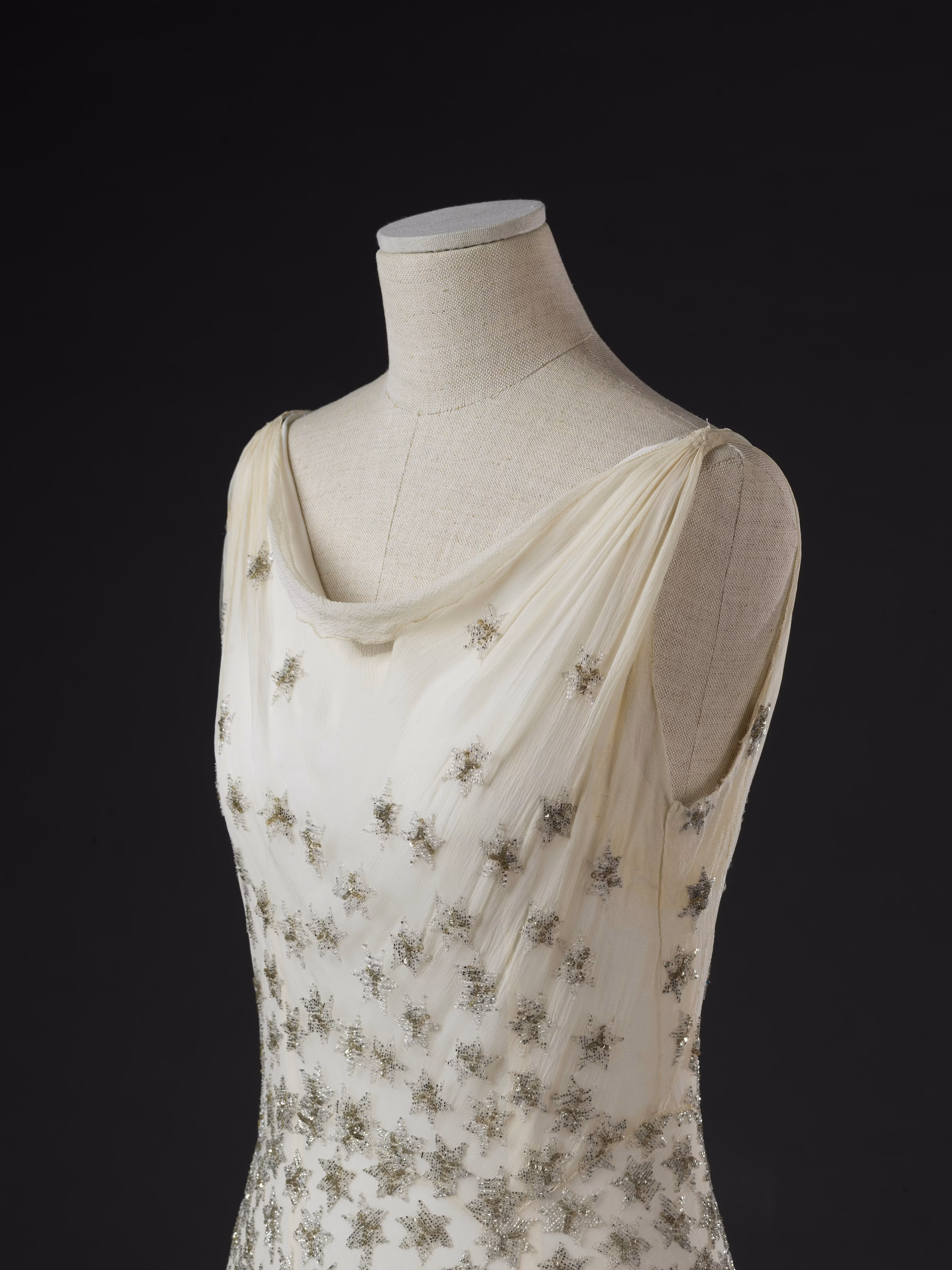Un elegante vestito bianco con spalline sottili, ricamato con elaborati motivi floreali in argento, esposto su un manichino contro uno sfondo nero.