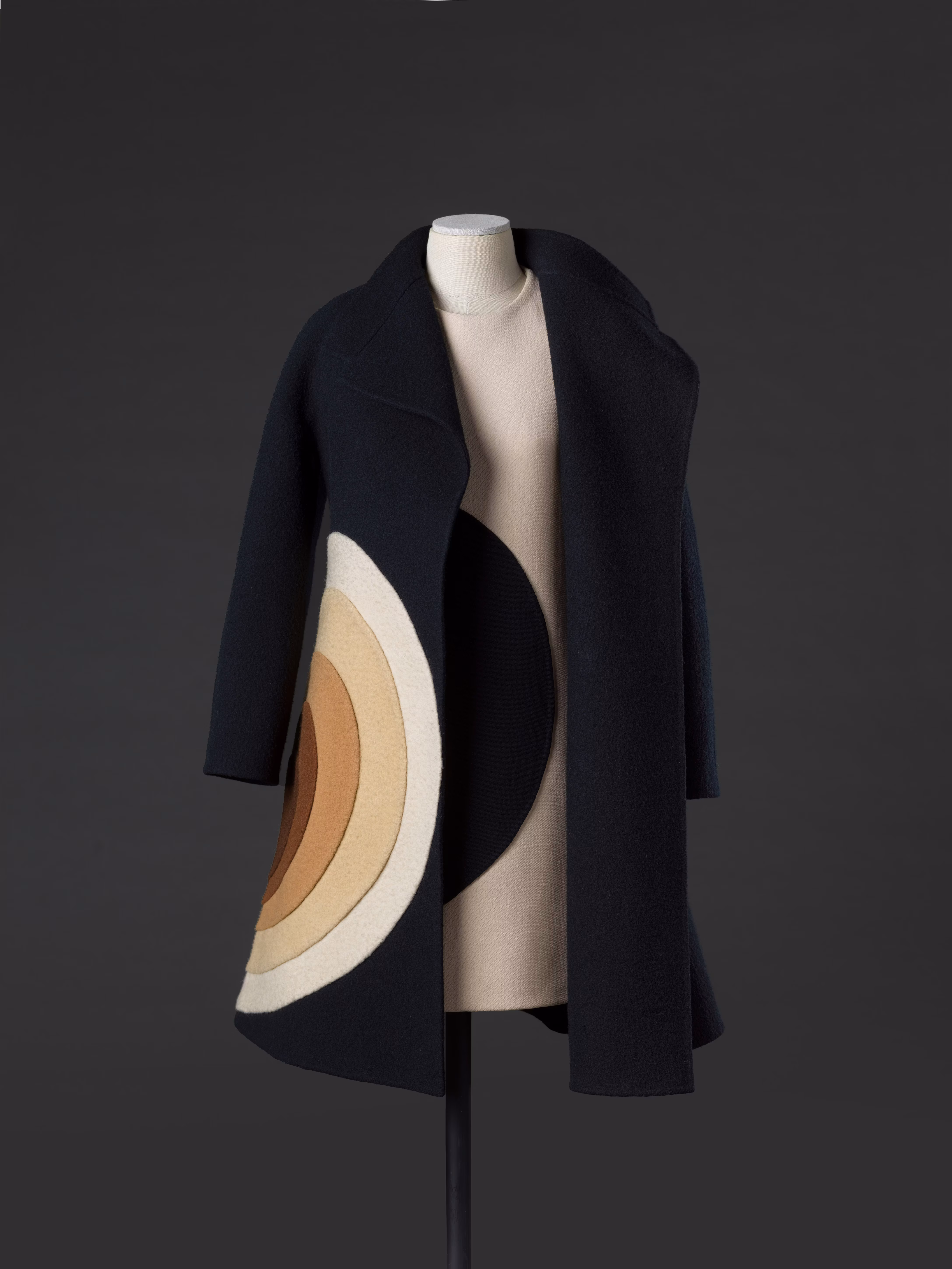 Un elegante cappotto nero su un manichino, caratterizzato da un ampio motivo circolare beige e marrone sul lato, posizionato su uno sfondo scuro.