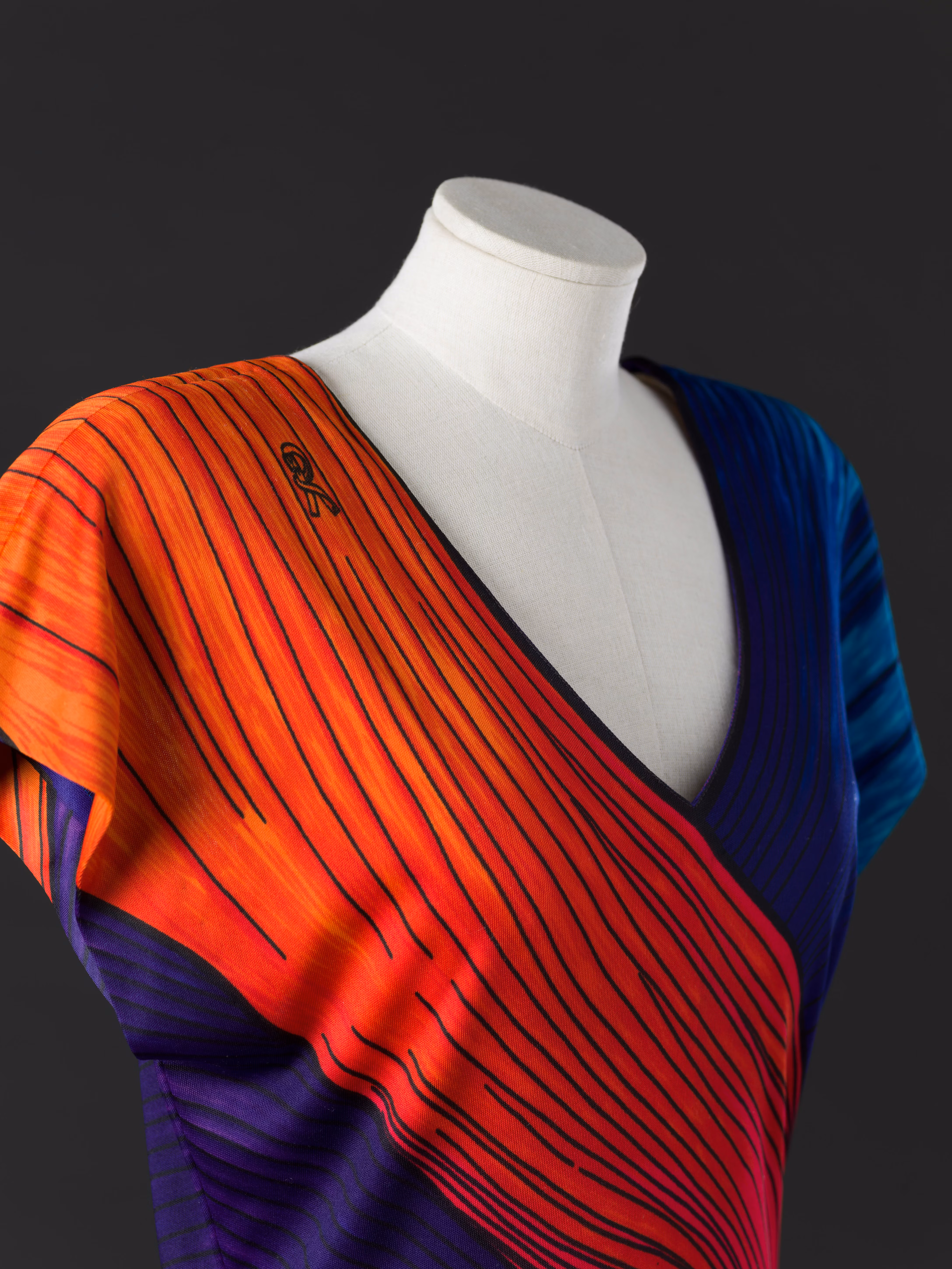 Un manichino indossa un vestito a righe con un design diagonale che combina tonalità vivaci di arancione, rosso, blu e viola, su sfondo nero.