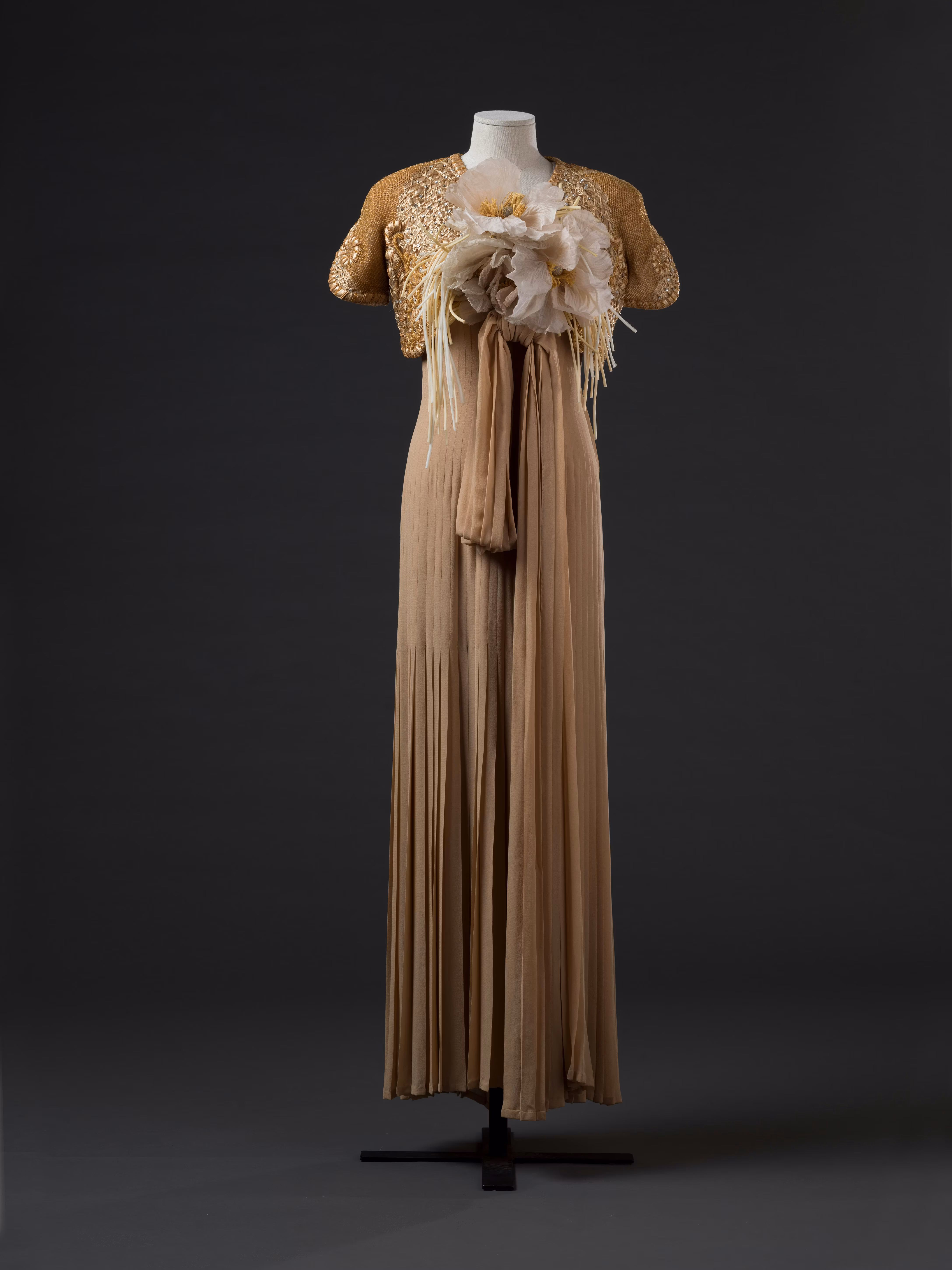 Abito elegante color crema con una gonna lunga e fluente e un bolero ricamato, arricchito da un grande dettaglio floreale sul busto, su uno sfondo scuro.