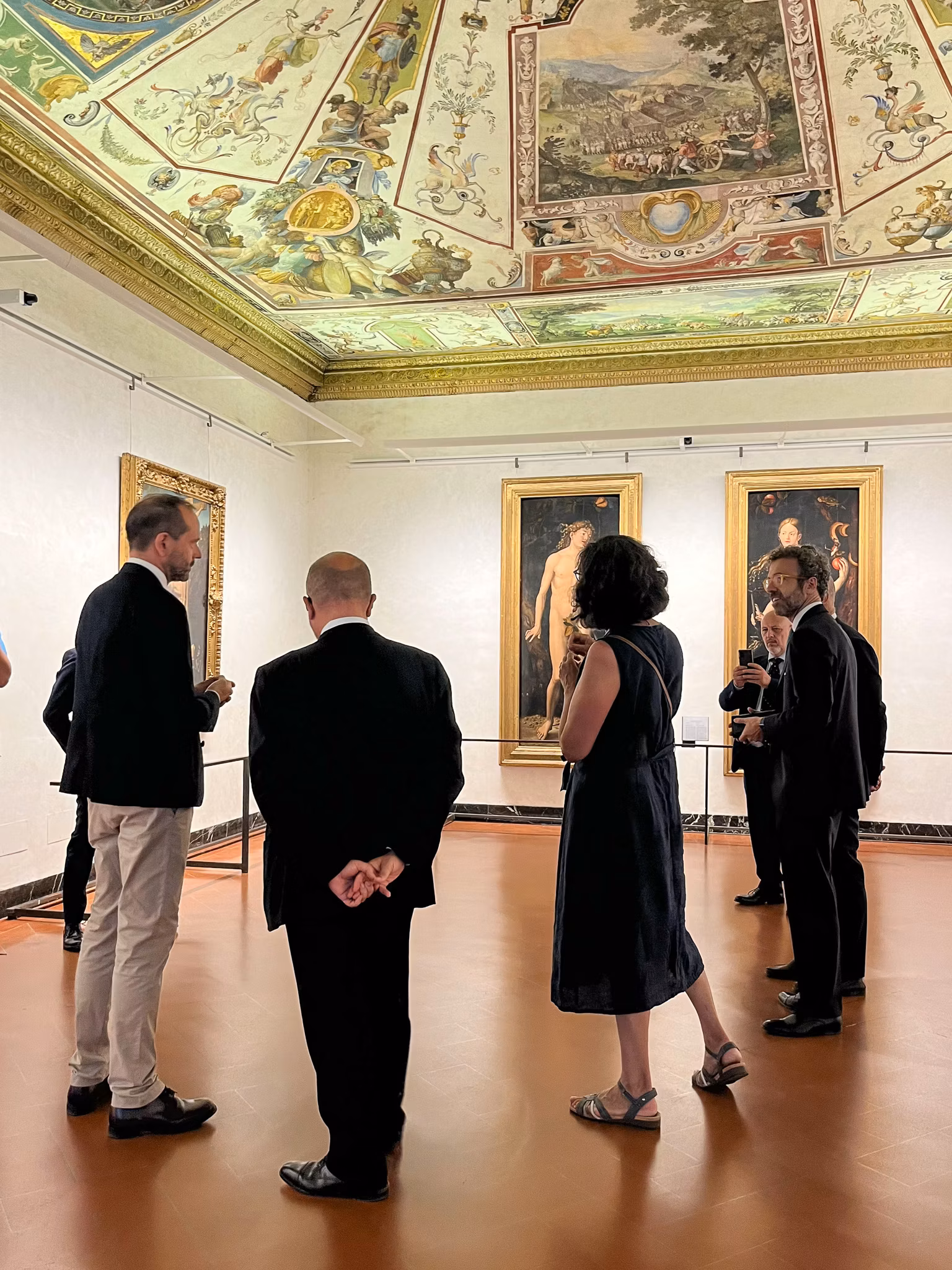 Un gruppo di persone osserva attentamente dipinti rinascimentali, incorniciati su pareti bianche, sotto un soffitto decorato riccamente con affreschi storici all'interno di una galleria d'arte.