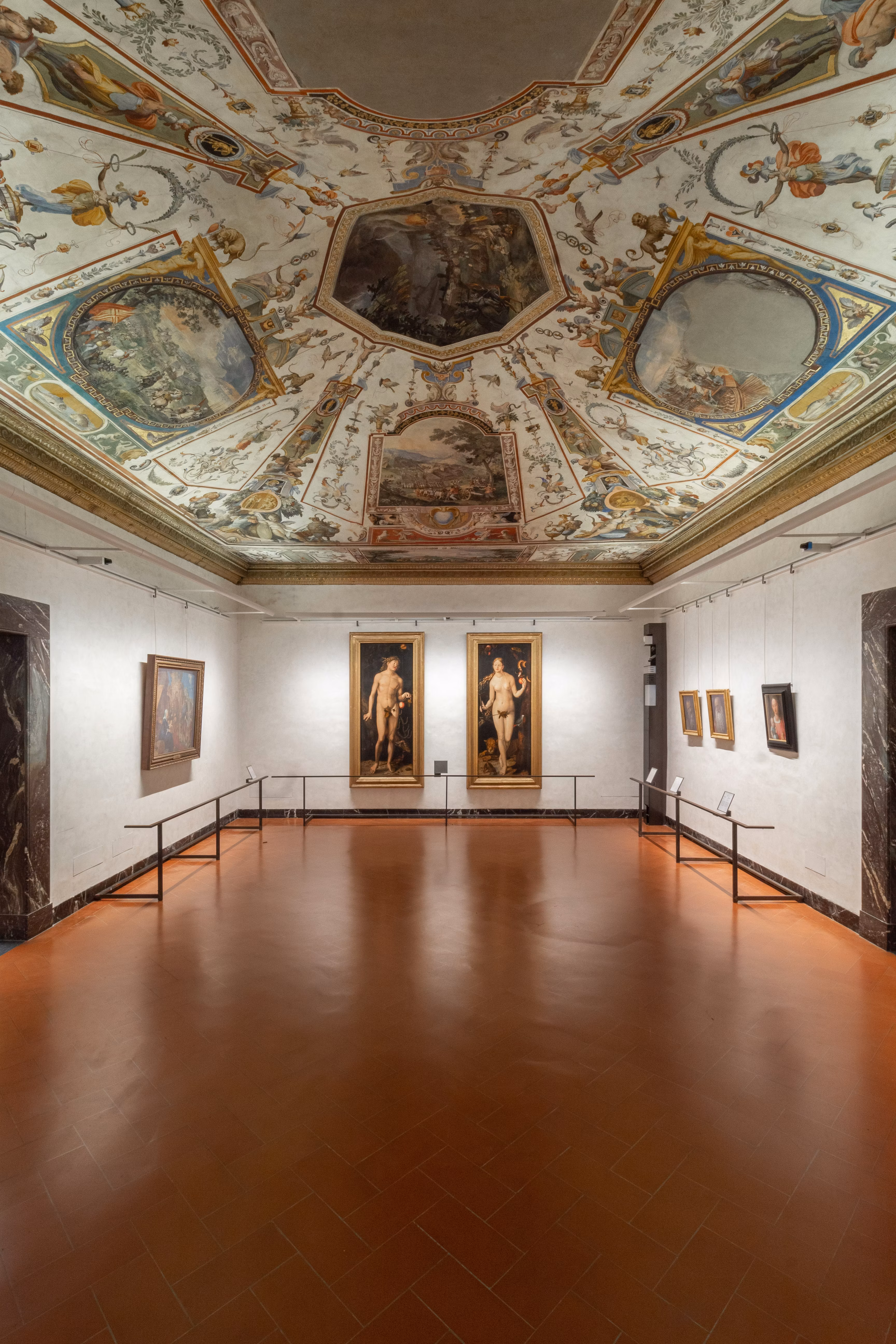 Una sala d'arte in un museo con pavimento in terracotta e pareti bianche, adornata da elaborati dipinti rinascimentali sul soffitto e ritratti incorniciati appesi lungo le pareti.