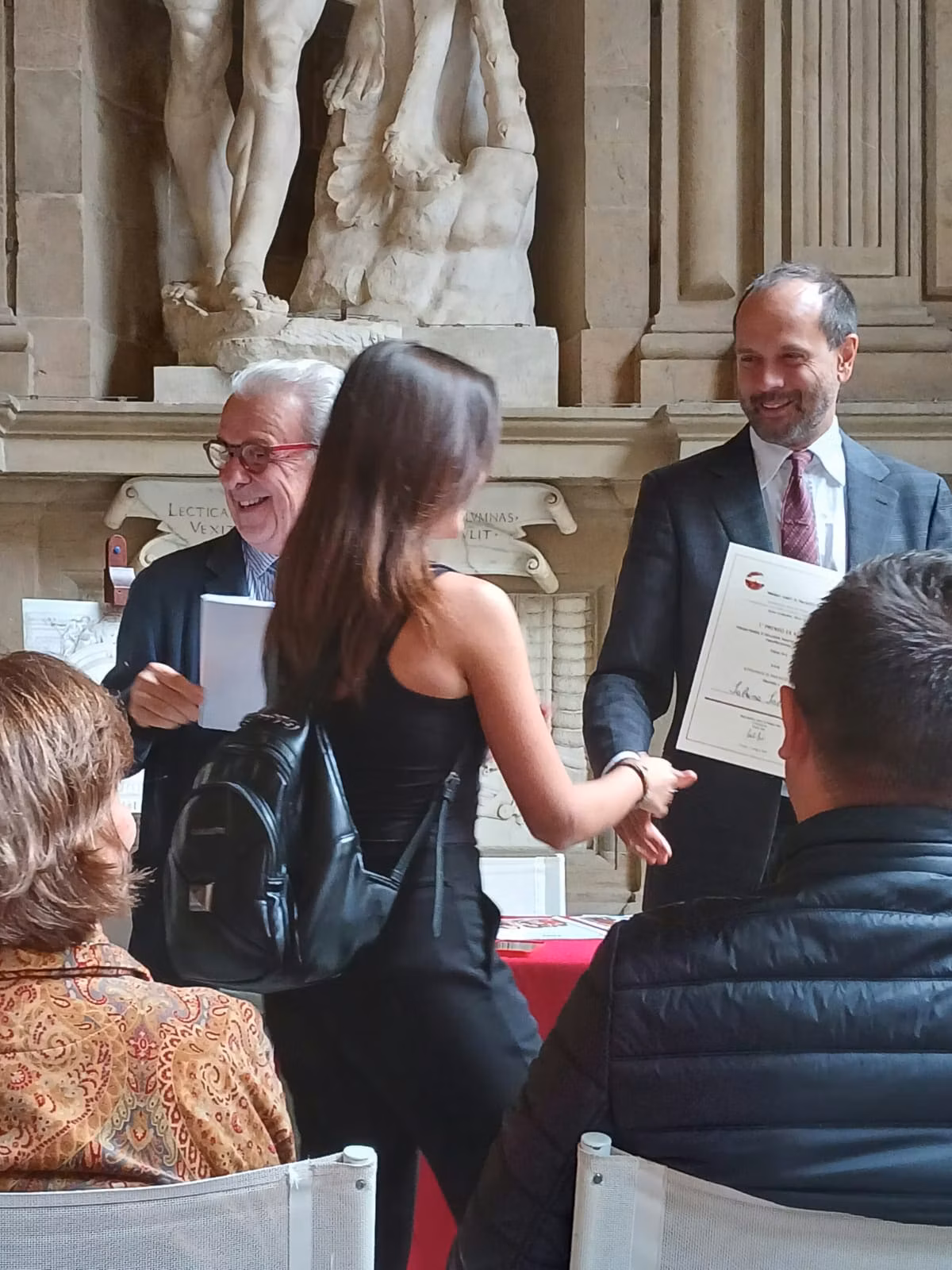 Una donna riceve un certificato in una cerimonia in un ambiente elegante con pareti in pietra e statue, sorridendo mentre stringe la mano a un uomo.