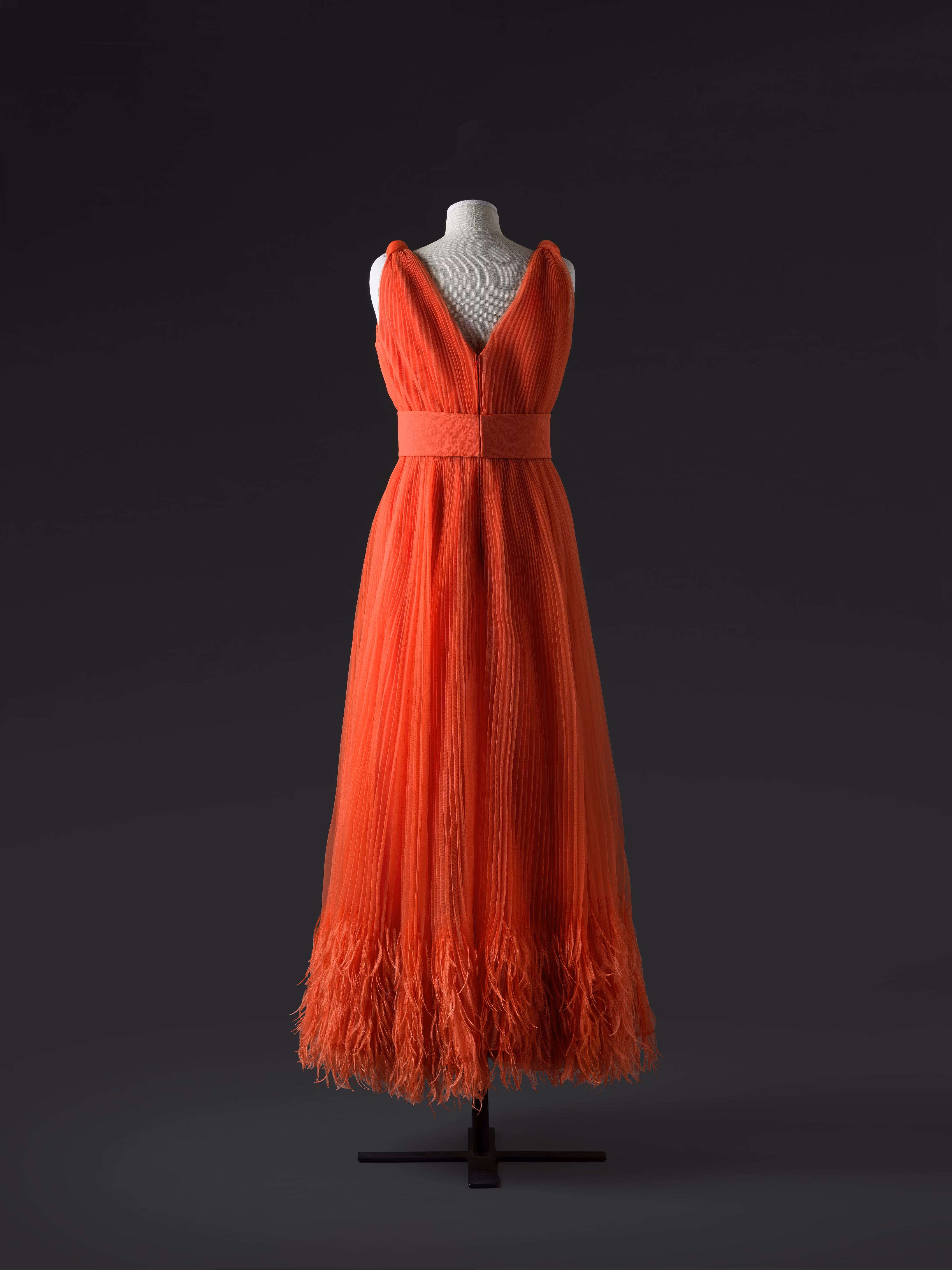 Un elegante manichino presenta un abito lungo rosso scuro, caratterizzato da un profondo scollo a V e una gonna plissettata che termina con dettagli di piume, il tutto su uno sfondo scuro che accentua il vibrante colore dell'abito.