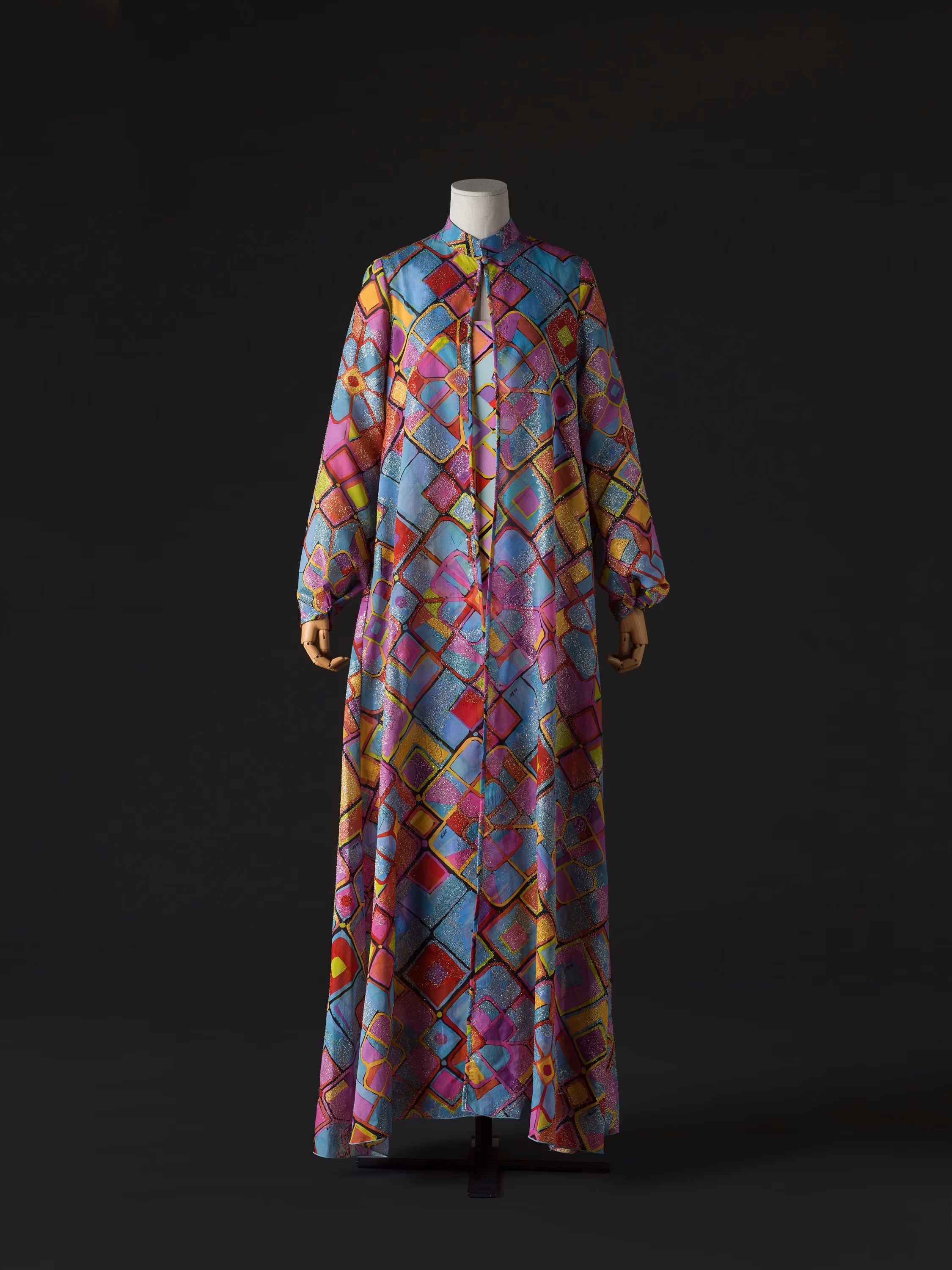 A mannequin displays a long, colorful geometric-patterned dress with vibrant blues, purples, and reds, set against a dark background.