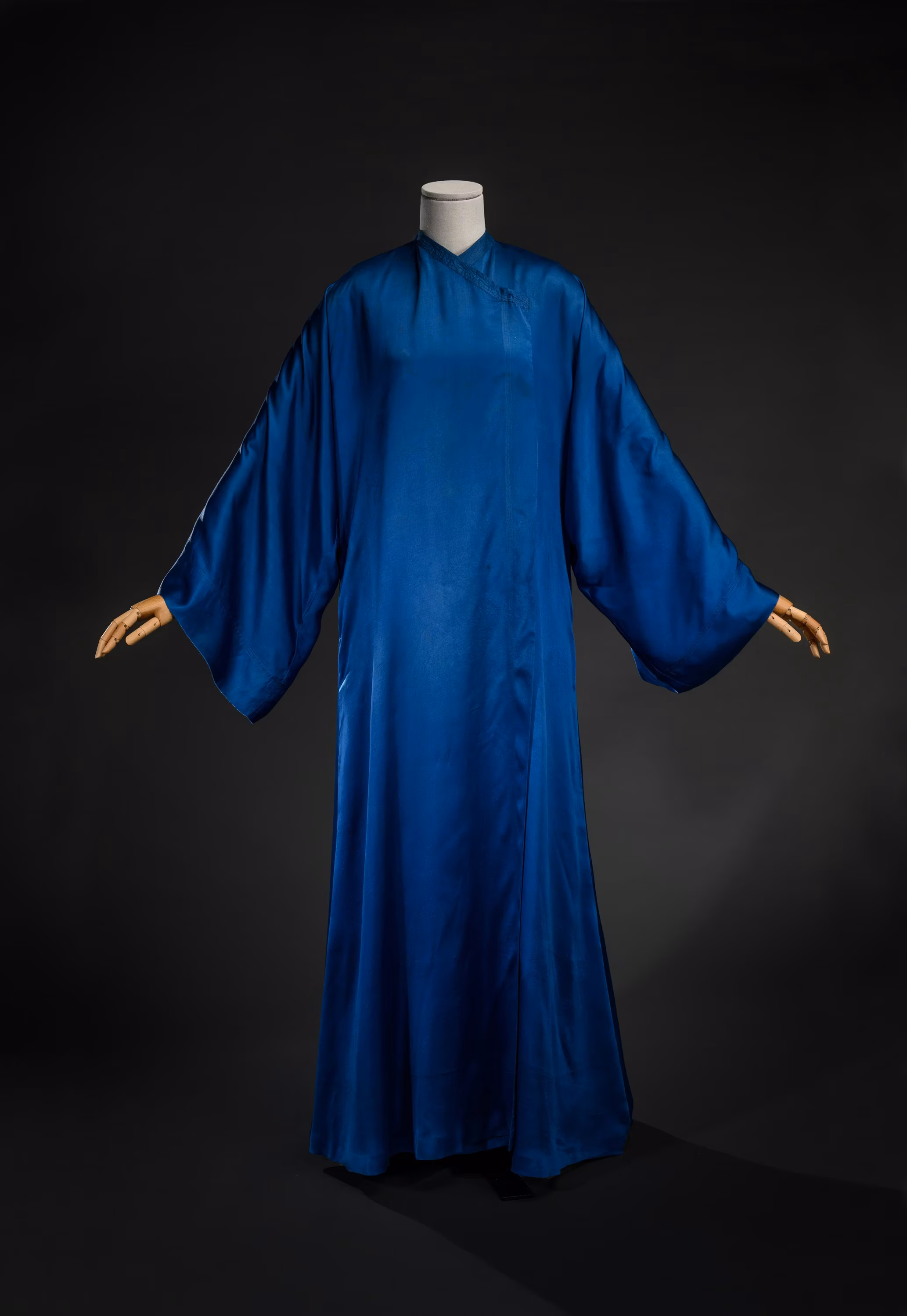 Un abito lungo in seta blu scuro con maniche larghe e fluenti, esposto su un manichino contro uno sfondo nero, evidenziando il design elegante e la qualità del tessuto.