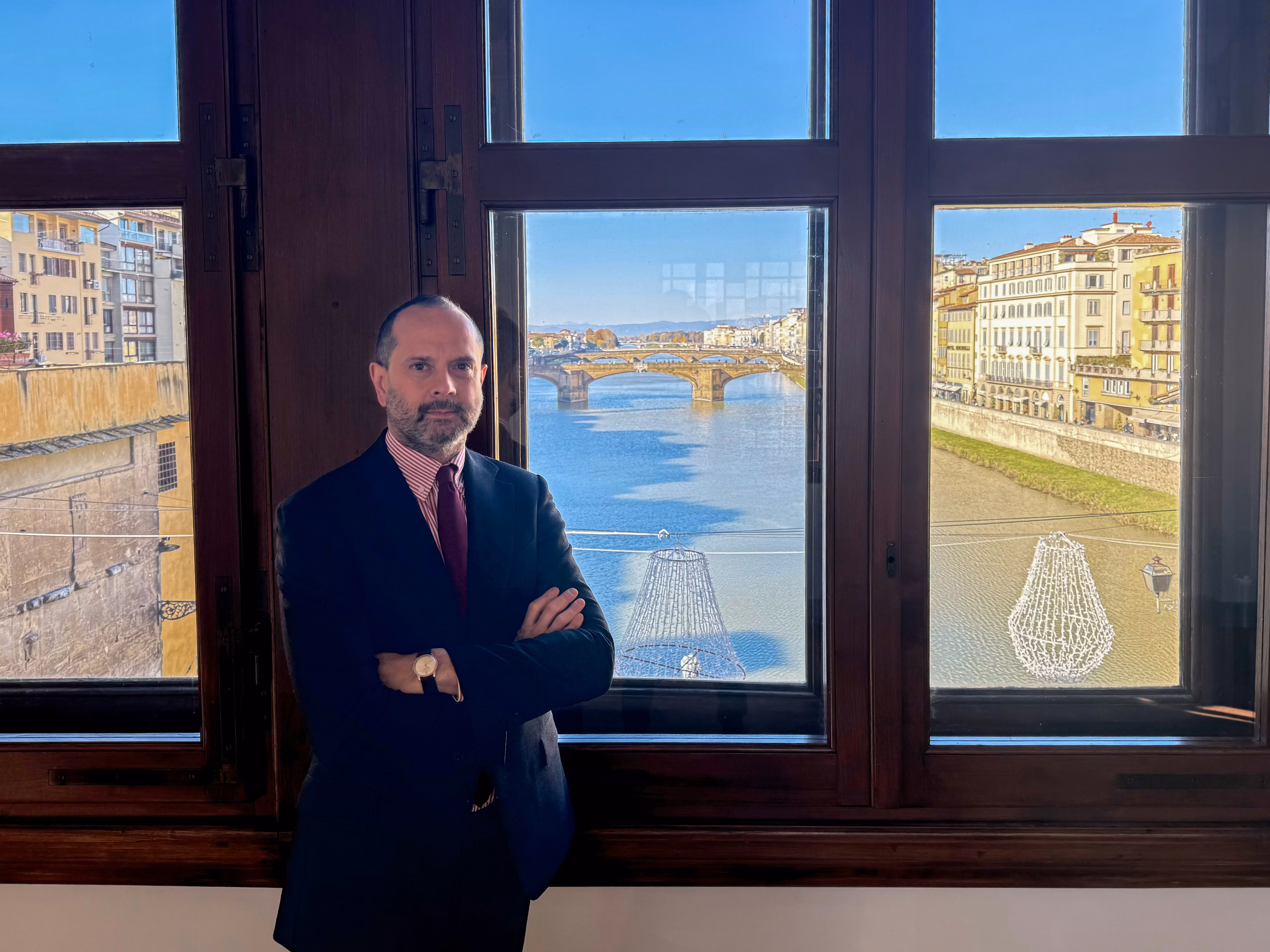 Un uomo in abito formale sta in piedi di fronte a una grande finestra, che offre una vista mozzafiato sul fiume Arno e il Ponte Vecchio a Firenze, in una splendida giornata di sole.