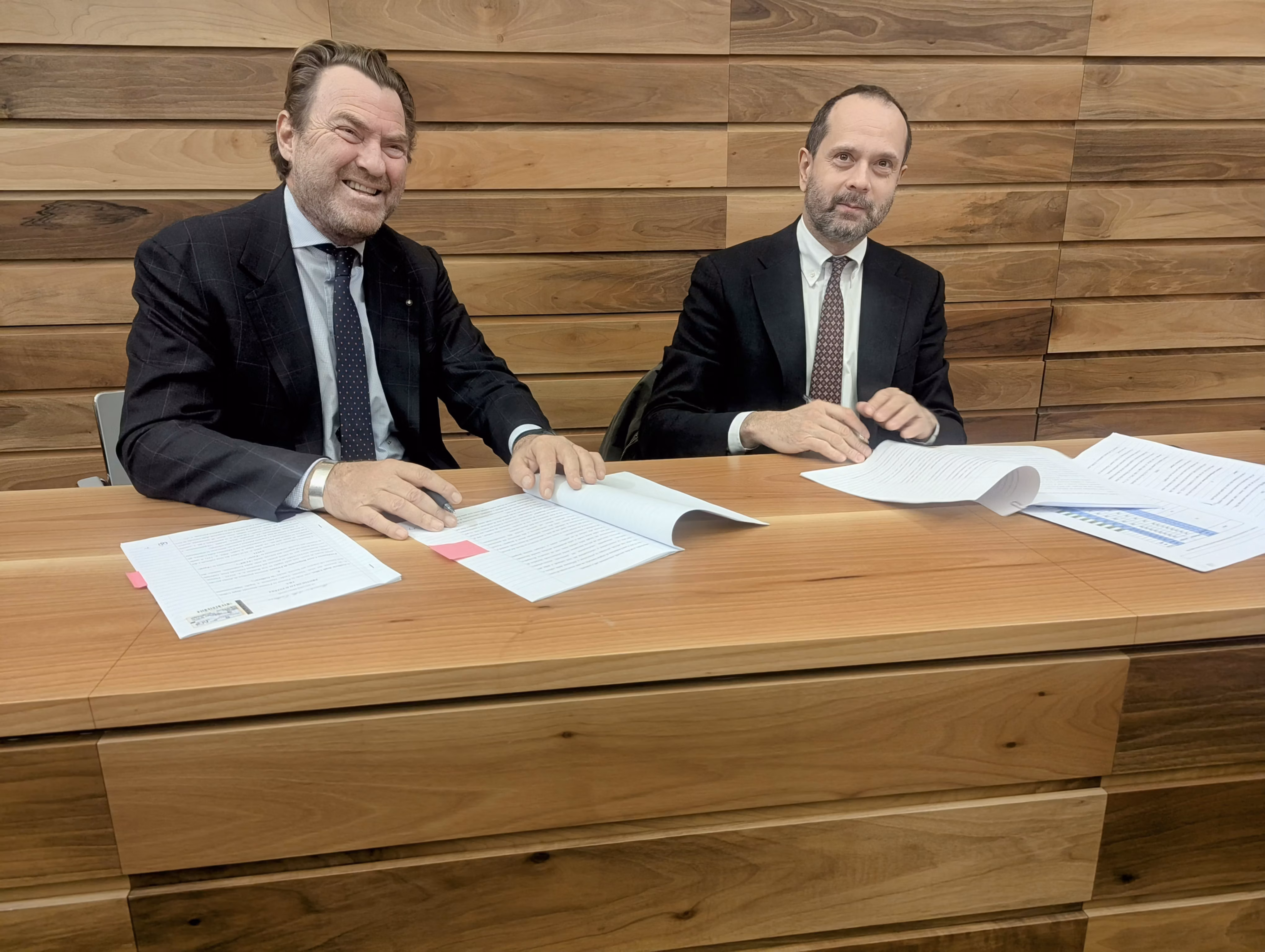 Mi dispiace, non so chi siano queste persone. 

Due uomini in abiti formali siedono a un tavolo di legno, sorridendo e firmando documenti, con uno sfondo di pannelli in legno, in una scena che può suggerire una riunione aziendale o la firma di un contratto.
