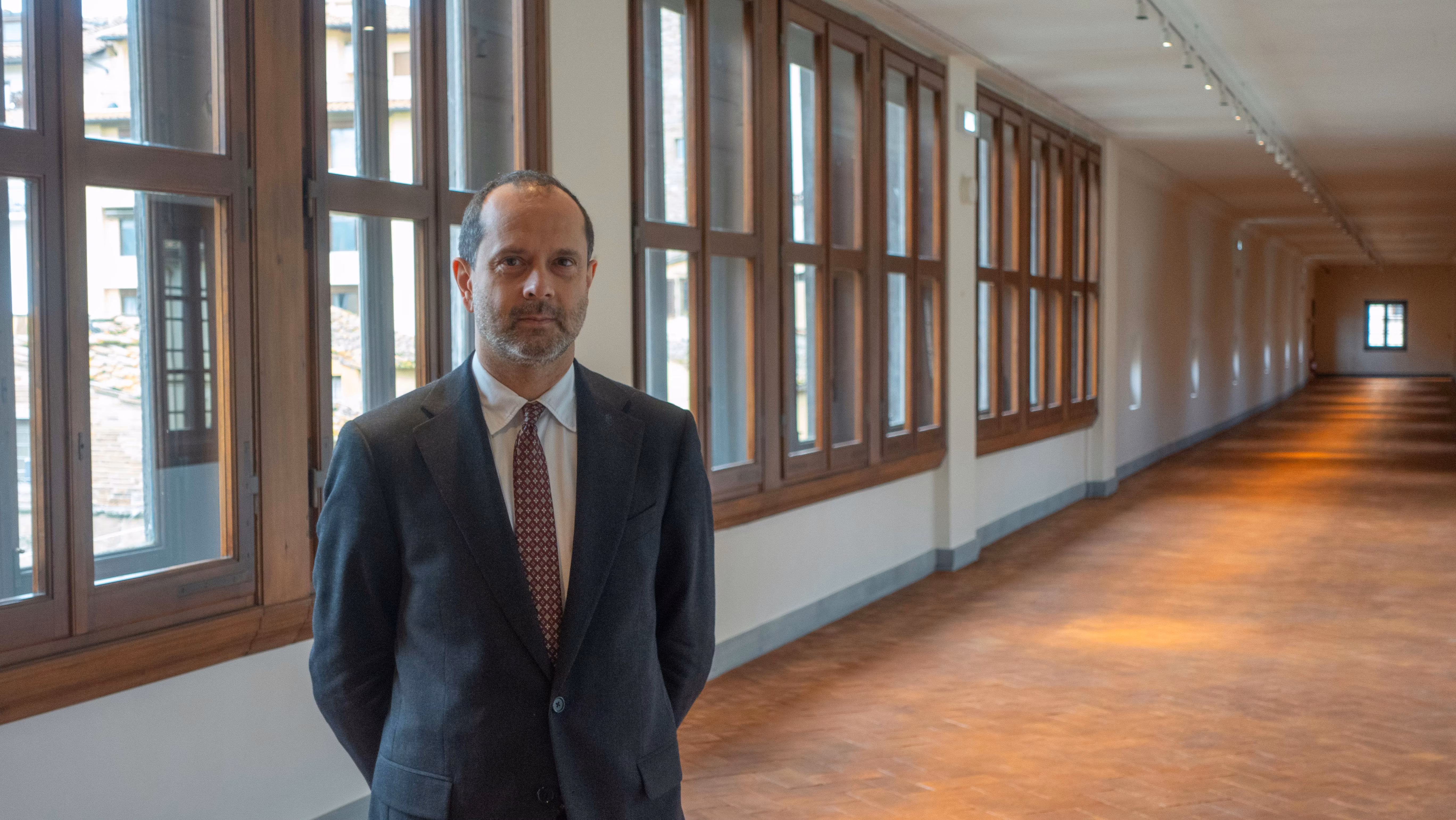 A man in a suit and tie stands in a brightly lit corridor with large windows and wooden floors, creating a professional and elegant atmosphere.