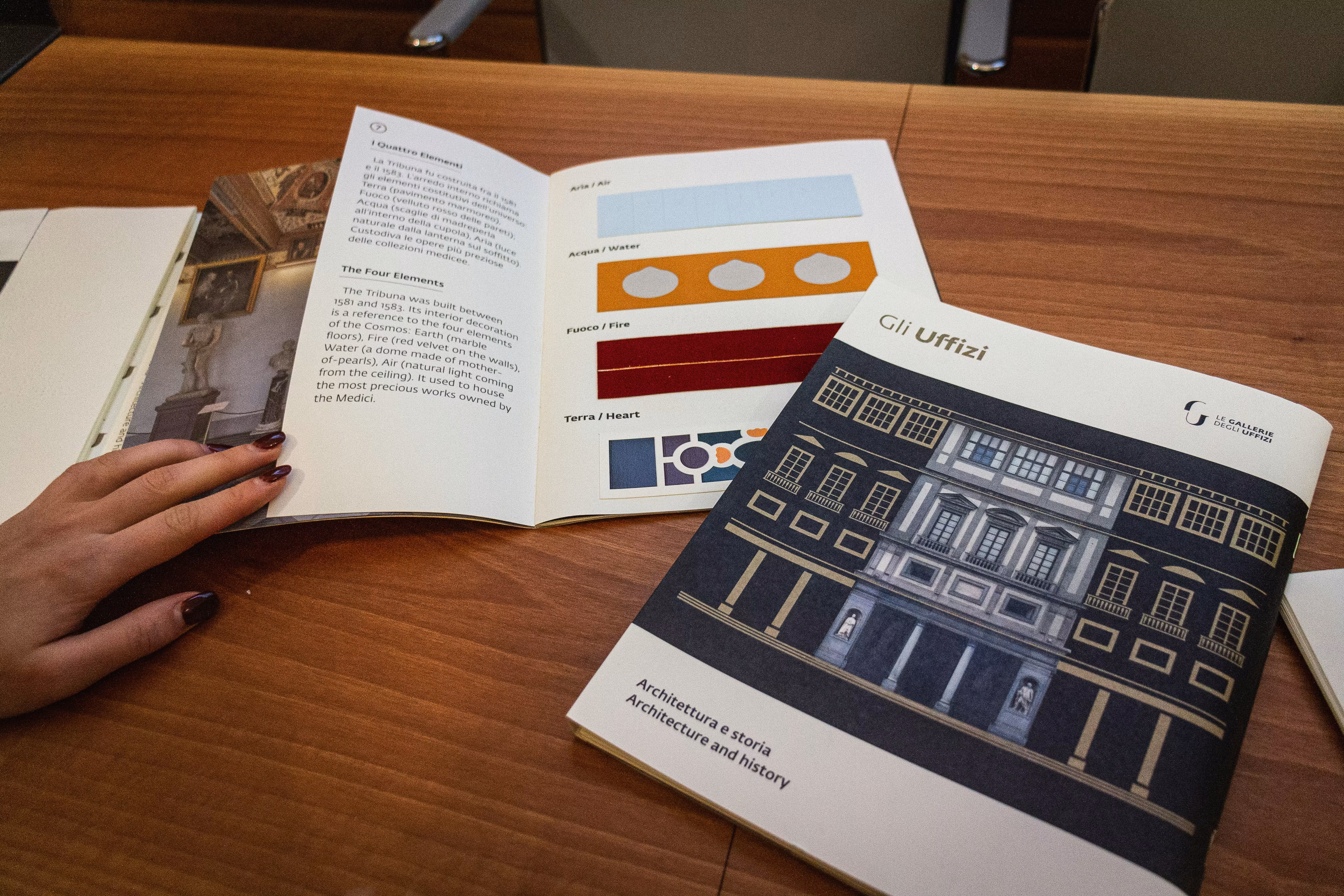 Una mano sfoglia un libro illustrativo sull'architettura del Giardino di Boboli, con accanto un catalogo aperto che mostra campioni di colori e fotografie di design.
