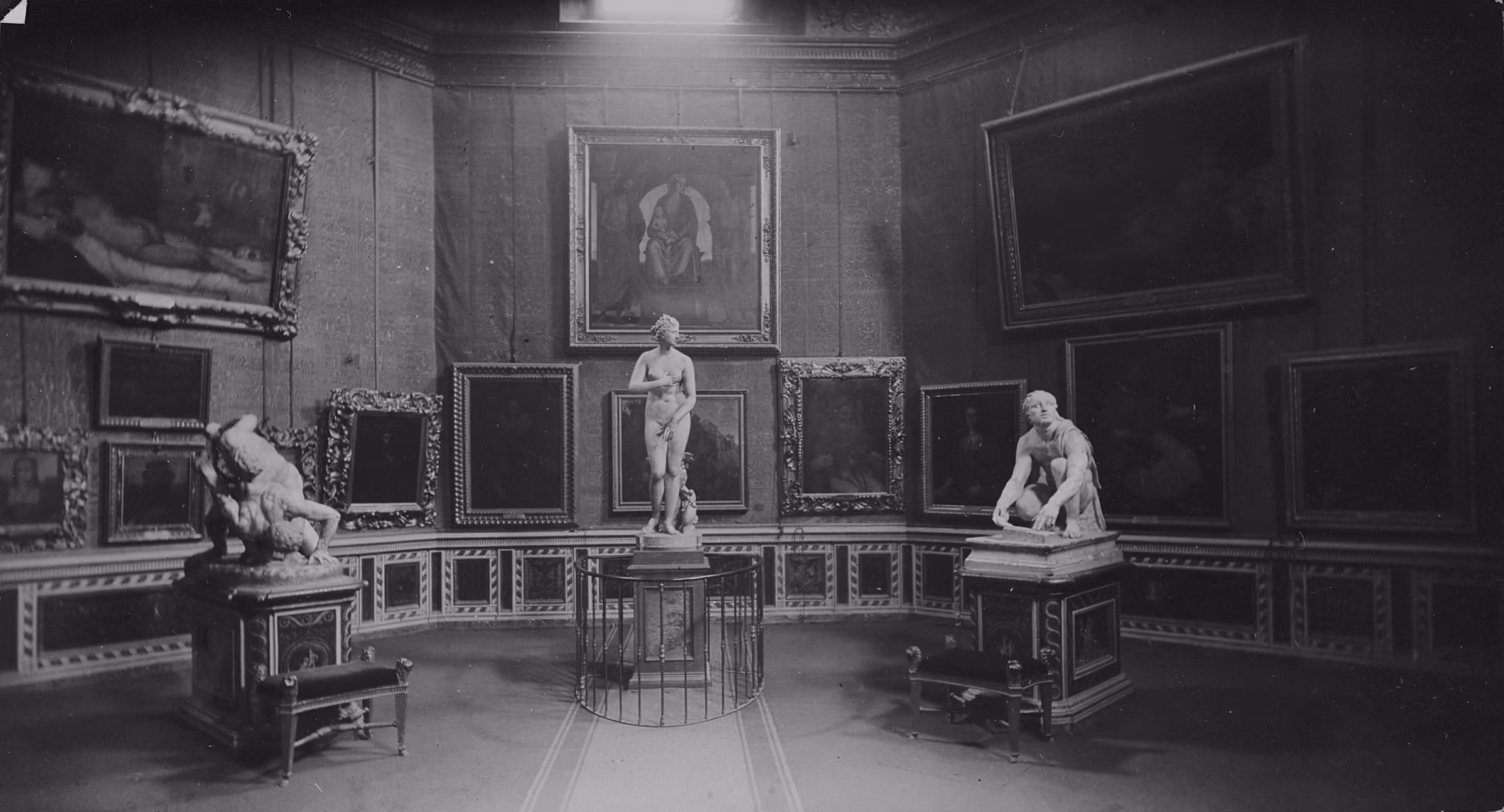 Una galleria d'arte con diverse opere appese alle pareti, tra cui dipinti incorniciati e due sculture classiche su piedistalli, illuminata dalla luce naturale che entra dall'alto.