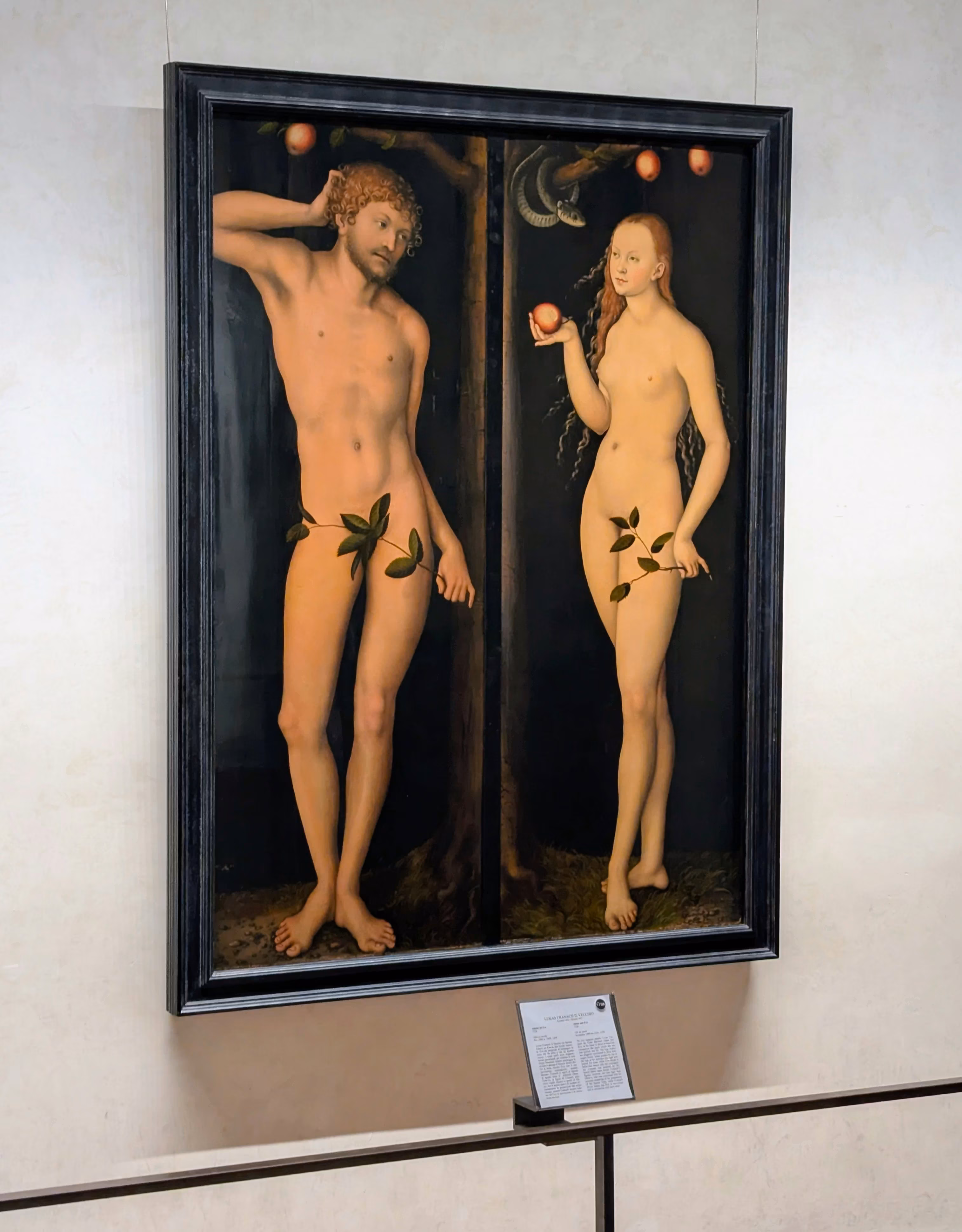 A framed painting depicting two nude figures, a man and a woman, reminiscent of Adam and Eve, standing in a dark, natural setting with strategically placed leaves for modesty; the woman holds an apple and branches extend above their heads.