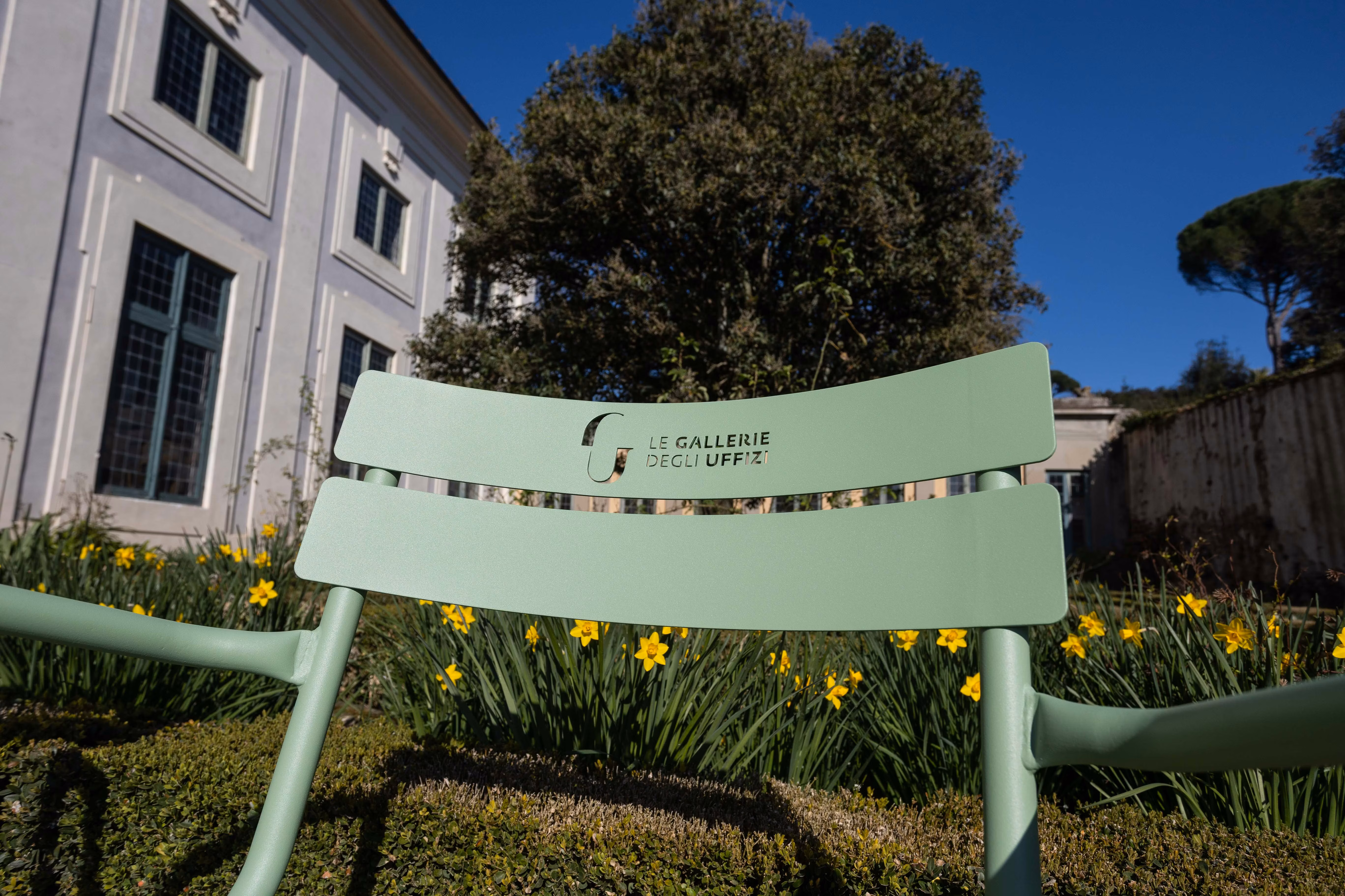 Una sedia verde chiaro con il logo "Le Gallerie degli Uffizi" è posizionata in un giardino fiorito con narcisi gialli, davanti a un edificio bianco con grandi finestre, sotto un cielo azzurro.