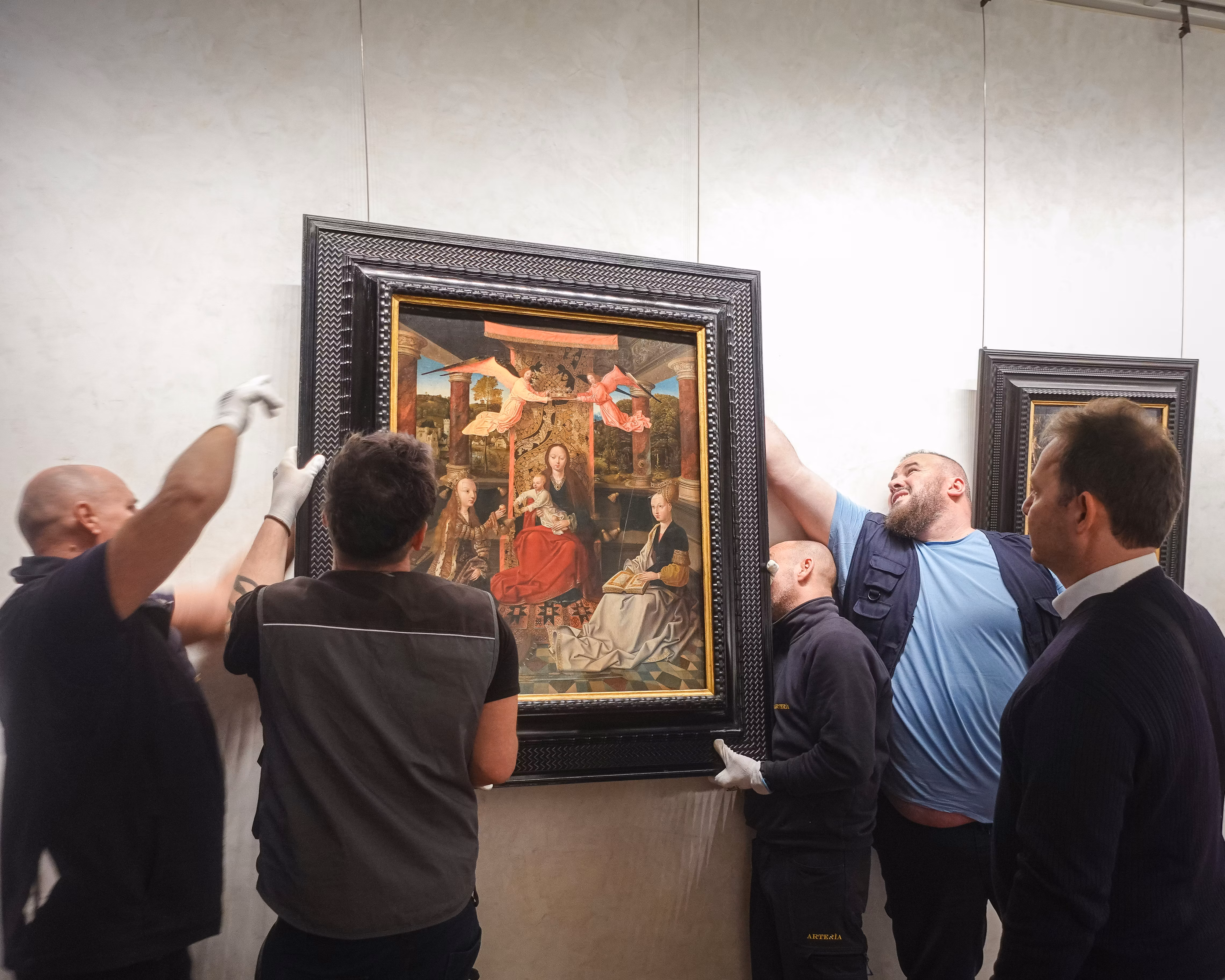 Cinque persone collaborano per appendere un grande dipinto in una cornice nera su una parete chiara, in una galleria d'arte.