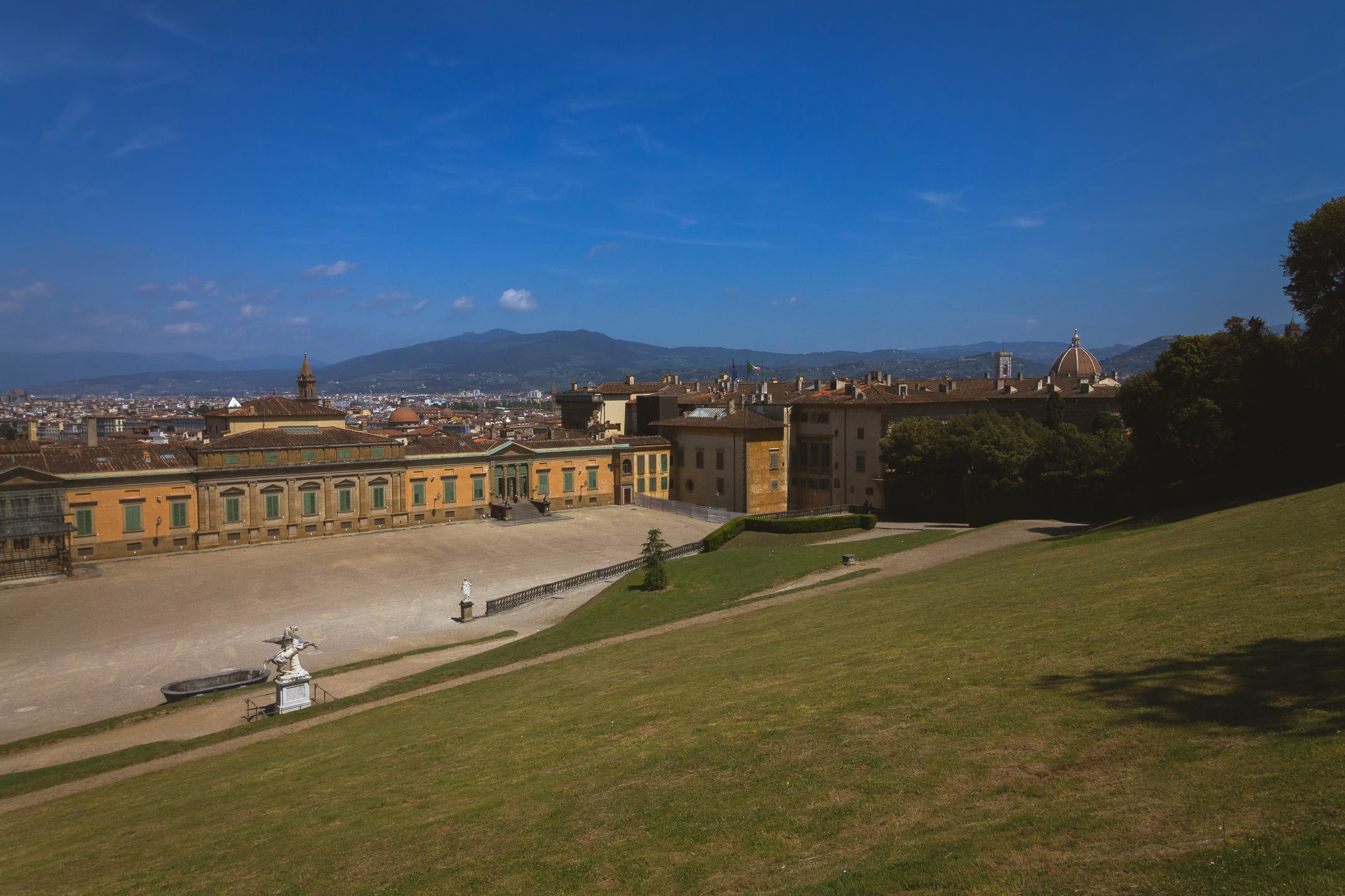 A panoramic view of the expansive Boboli Gardens in Florence features lush green lawns, historical architecture, and the distant silhouette of the Florence Cathedral under a clear blue sky.