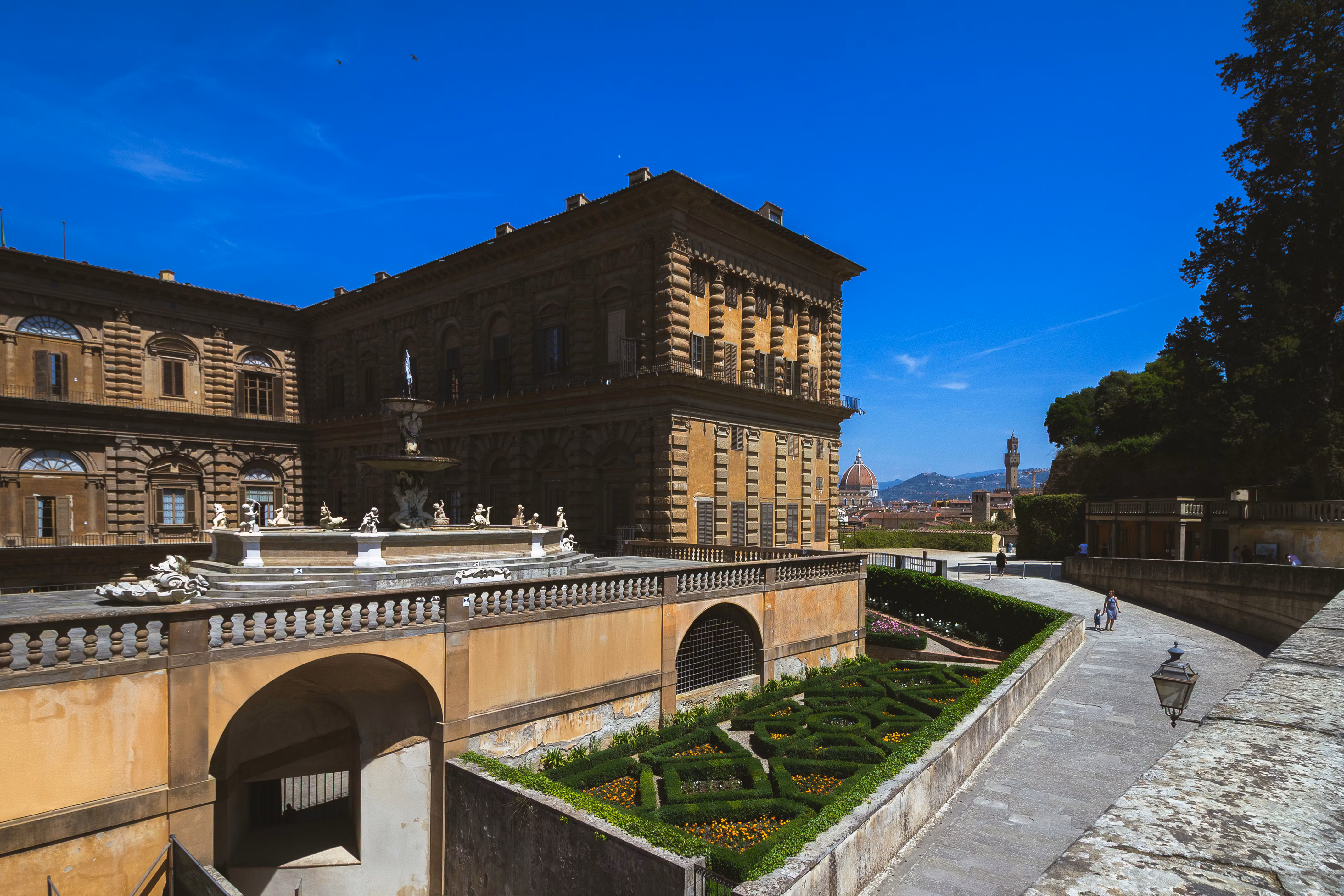 A historic building with ornate architectural details stands against a bright blue sky, overlooking a meticulously manicured garden featuring geometrically shaped hedges, in Florence.