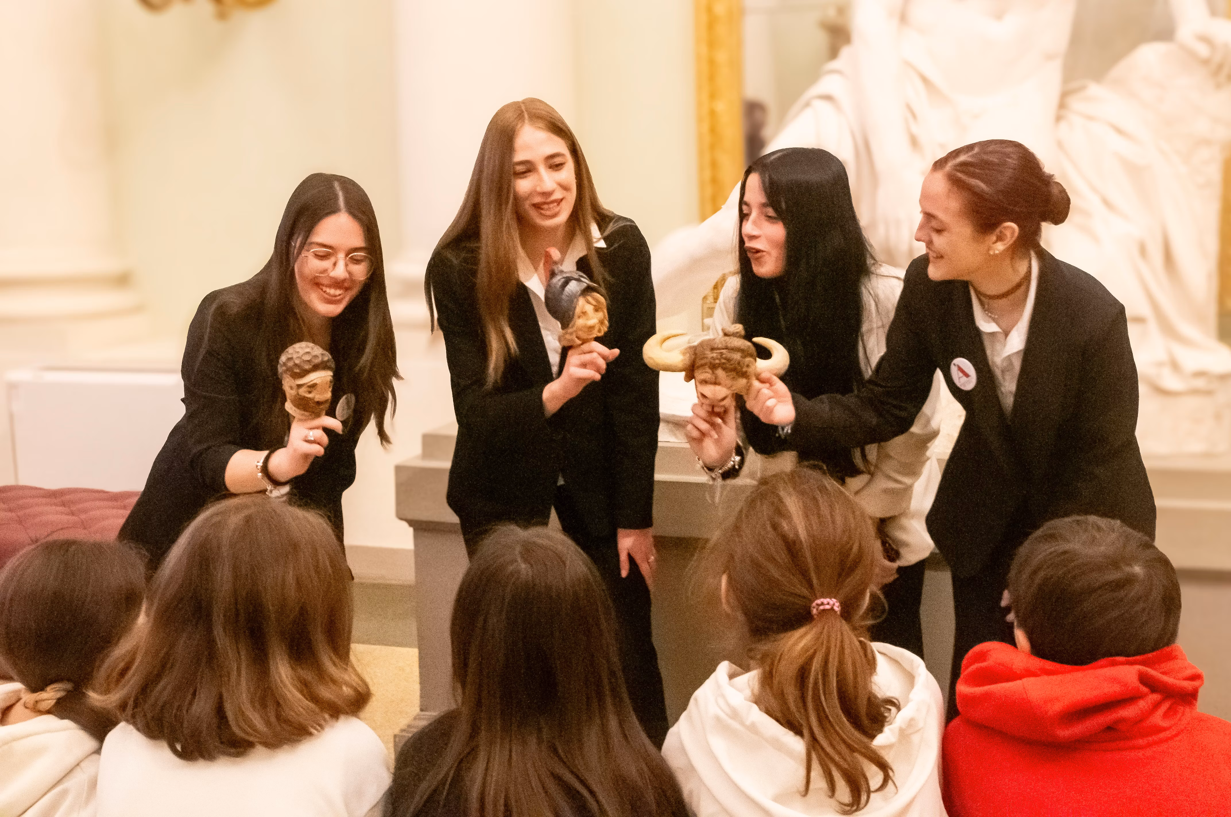 Un gruppo di quattro giovani donne in abiti formali mostra maschere teatrali a un gruppo di bambini seduti, all'interno di un museo con statue e colonne bianche nello sfondo.